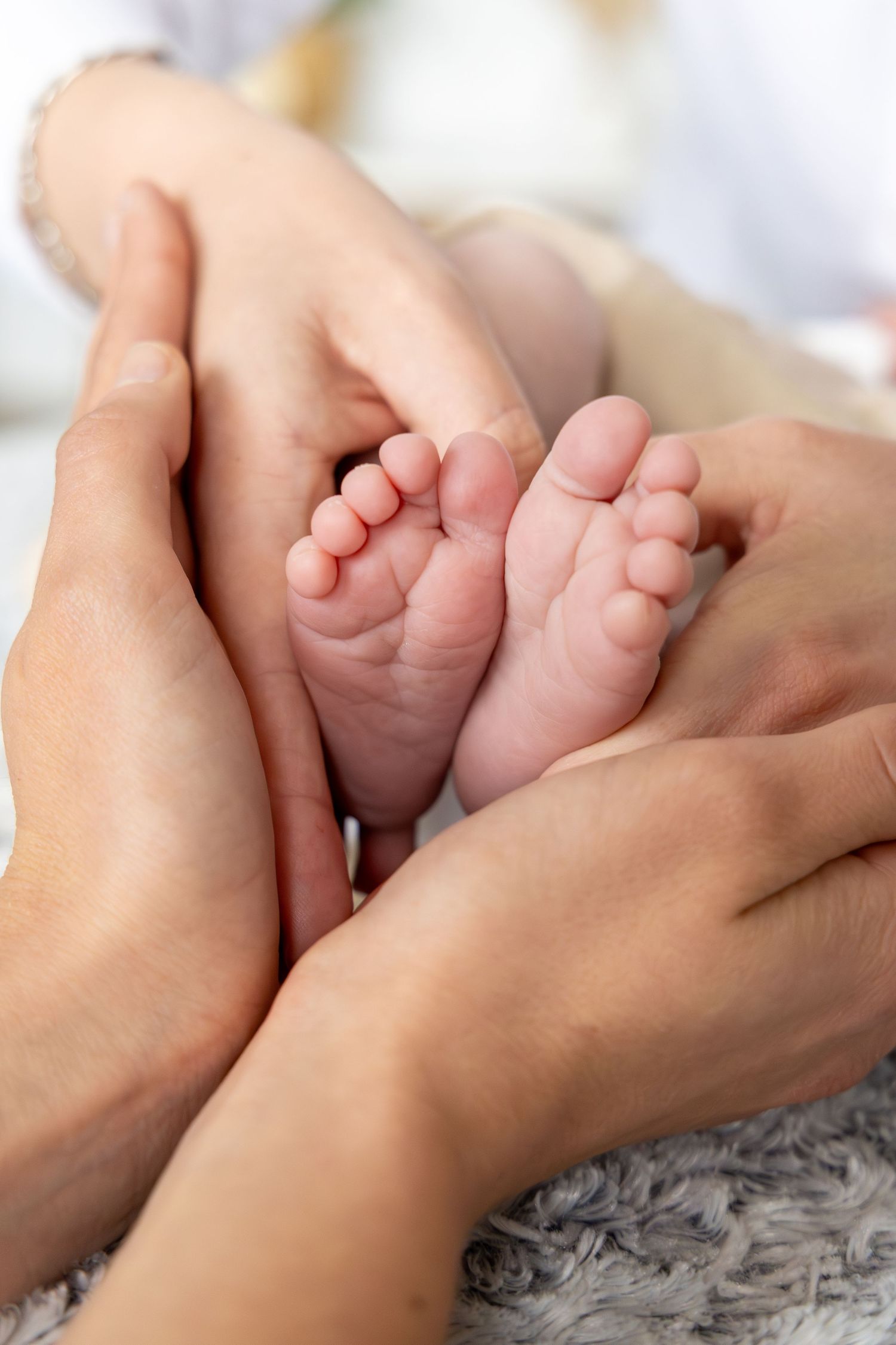 Adult hands gently cradling tiny newborn baby feet on a soft gray blanket, symbolizing parental love and care.