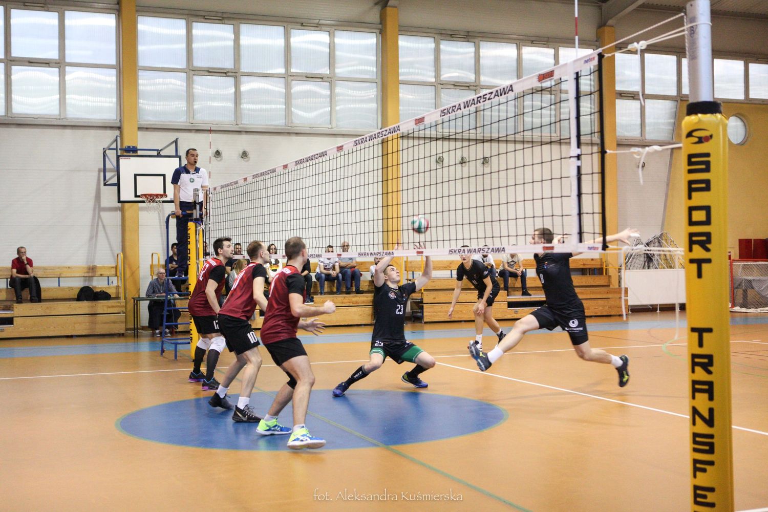 Men playing competitive indoor volleyball match in gymnasium with yellow net post.