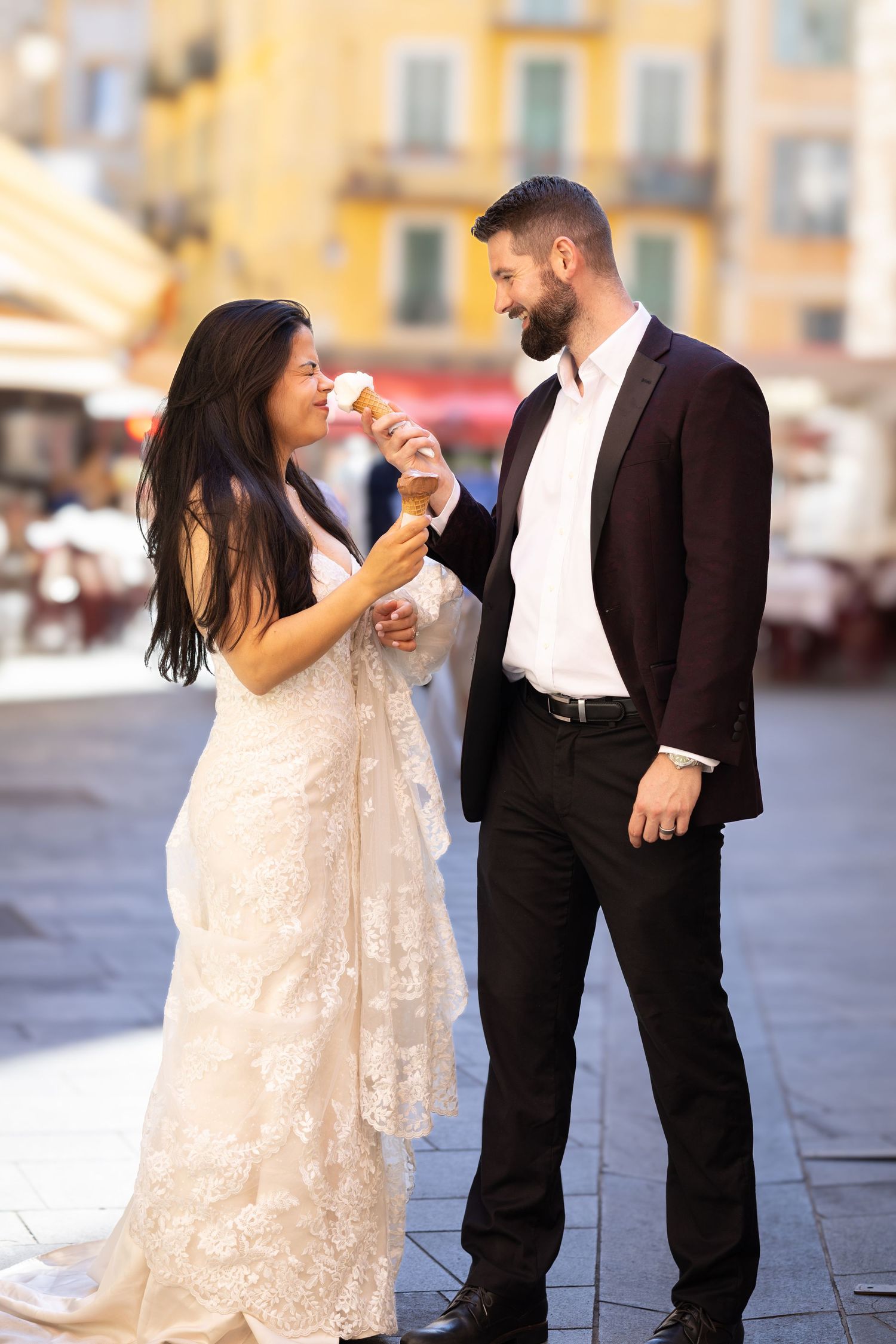 Bride in lace gown eating ice cream cone with groom in black suit on a sunny town square.