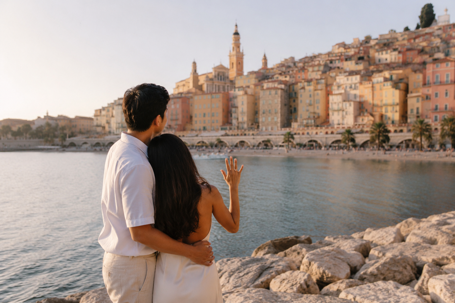 Couple embracing on rocky shore admiring colorful coastal town buildings at golden hour sunset.