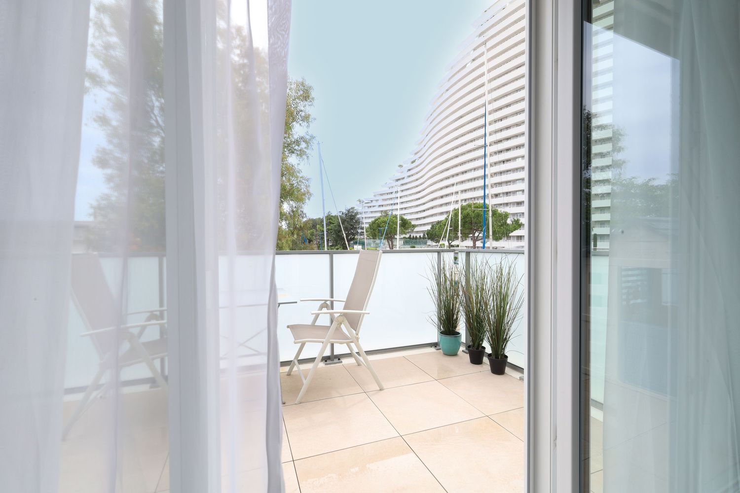 Sunny balcony with white chairs and potted plants overlooking a modern white building exterior.