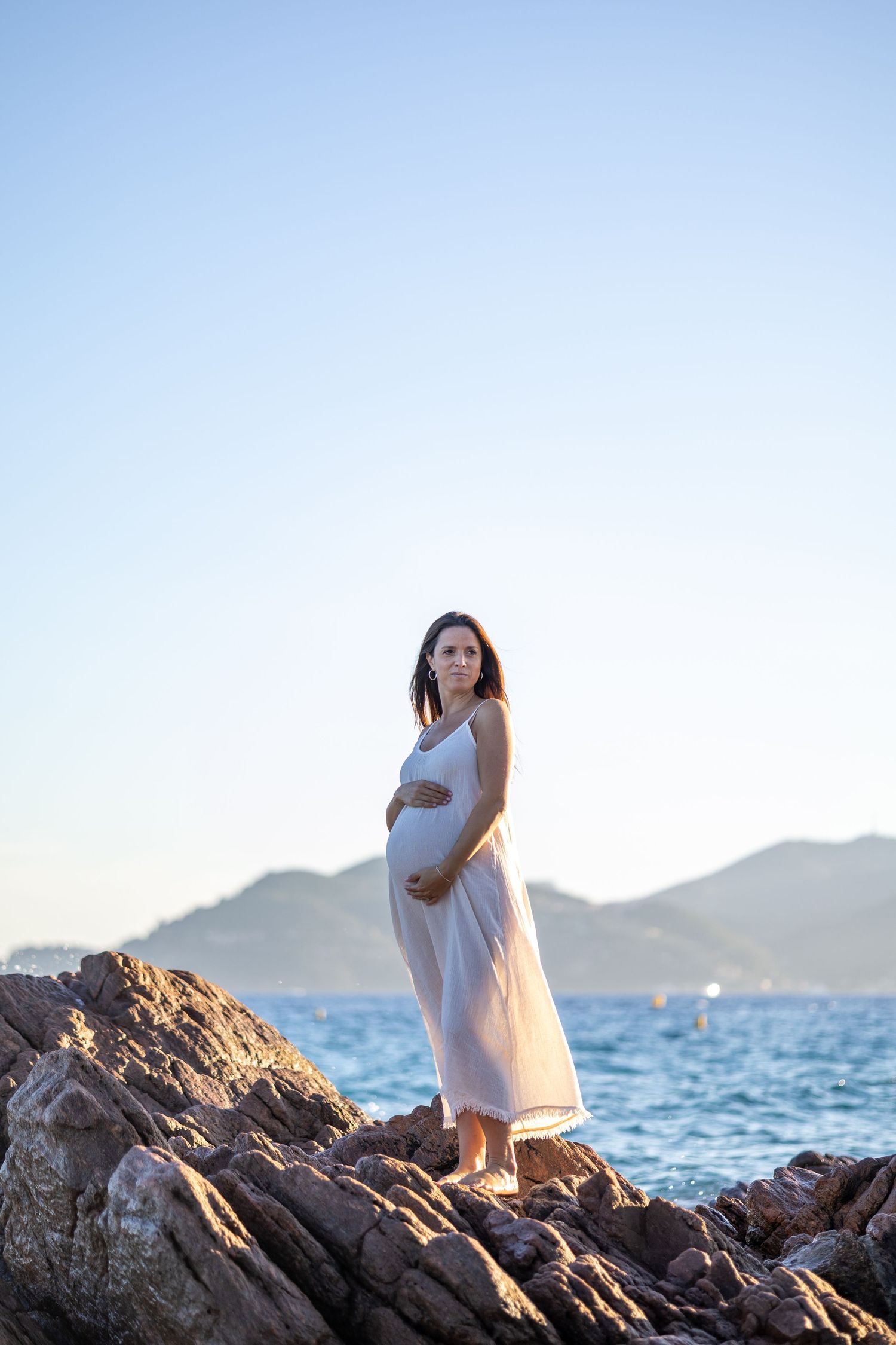A pregnant woman in a white dress stands on coastal rocks, cradling her belly at sunset.