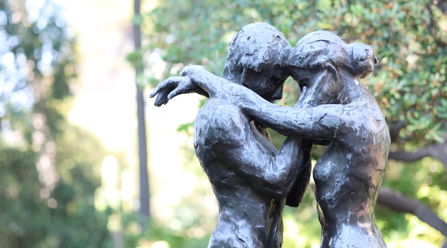 Bronze sculpture of two intertwined figures embracing outdoors, surrounded by lush green trees in a garden setting.