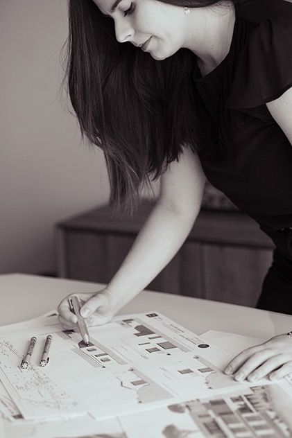 Woman in black top leaning over desk reviewing architectural drawings with pen in hand.