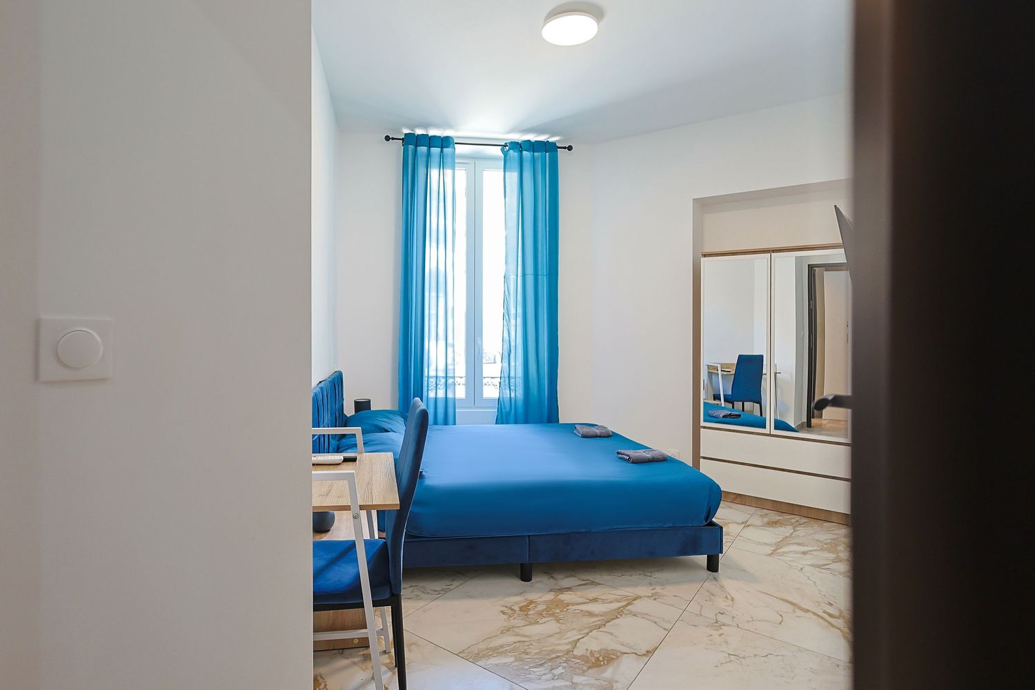 Bright hotel room with blue curtains, blue sofa bed, and marble flooring.