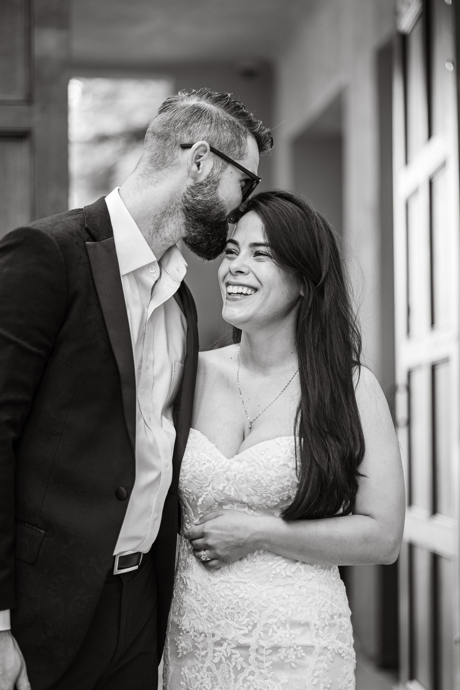 Black and white portrait of laughing bride with groom whispering in her ear.