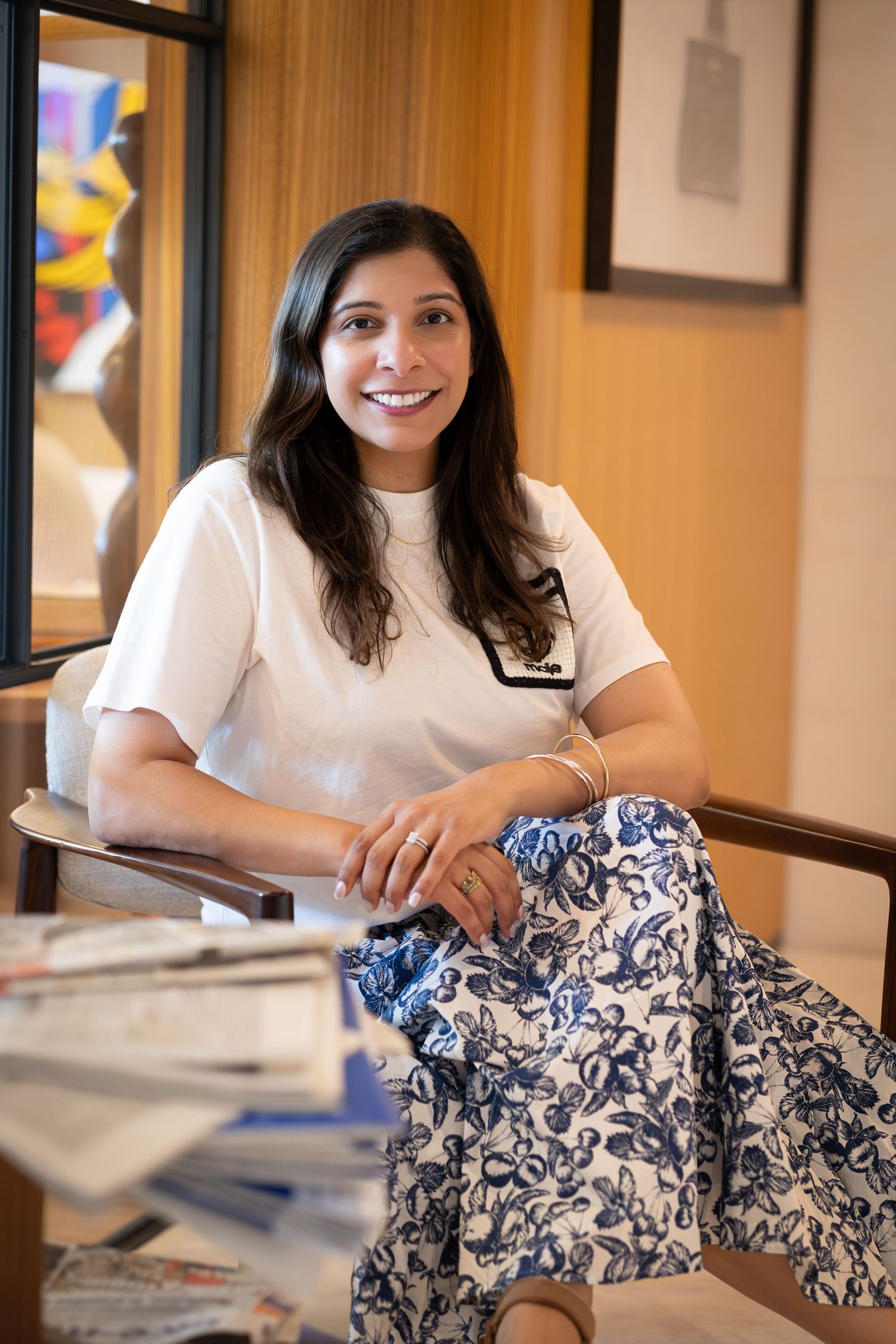Smiling woman in white top and floral skirt sitting relaxed in a modern office setting with artwork in background.