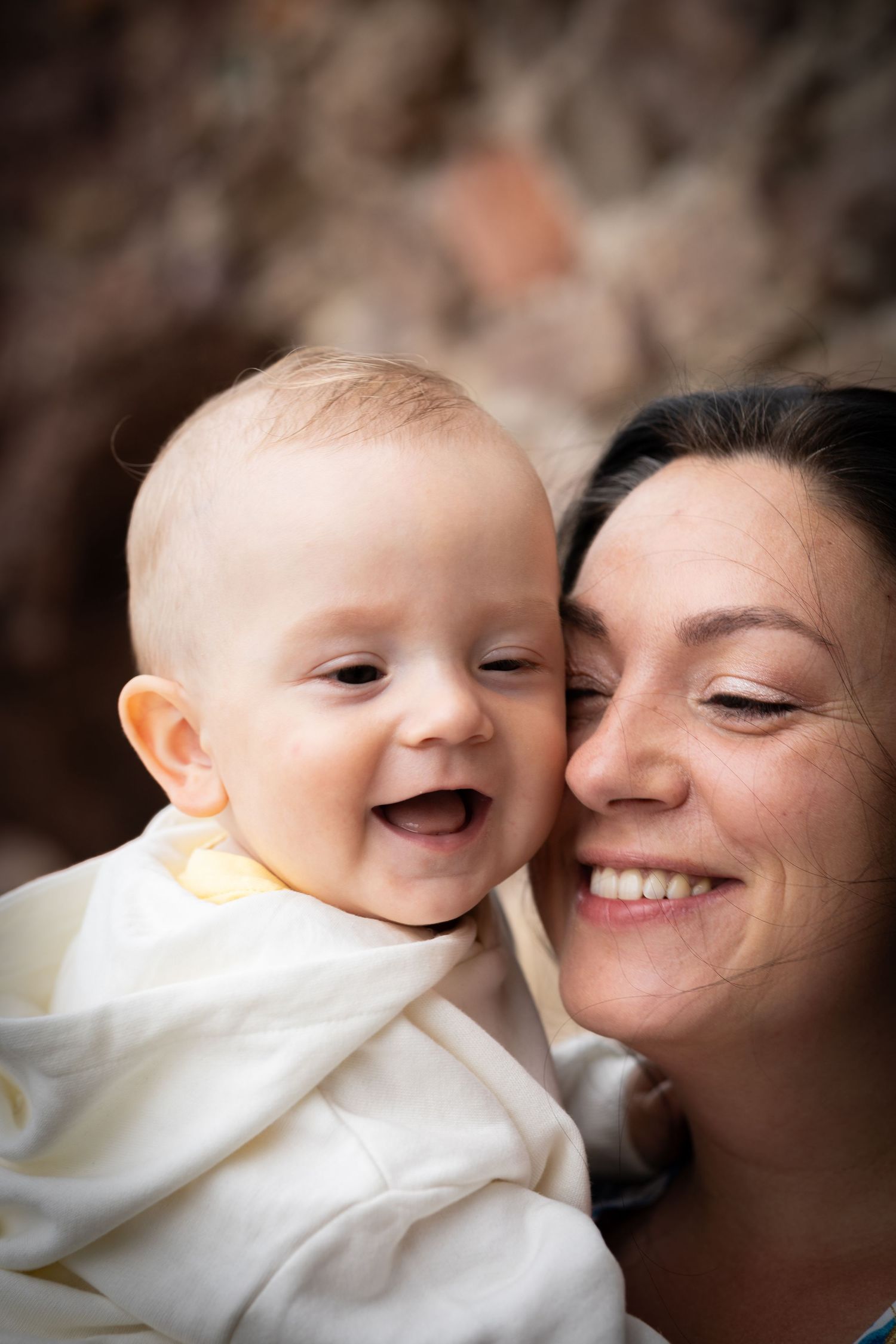 Joyful baby laughing cheek-to-cheek with a smiling dark-haired mother against a rocky background.
