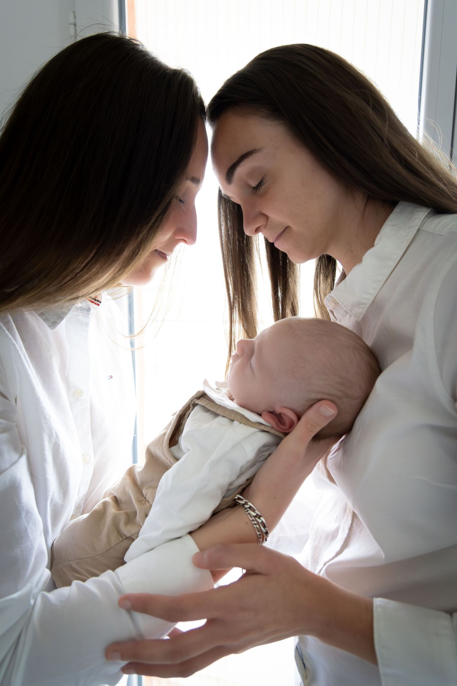 Two women in white shirts tenderly holding a sleeping newborn baby together.
