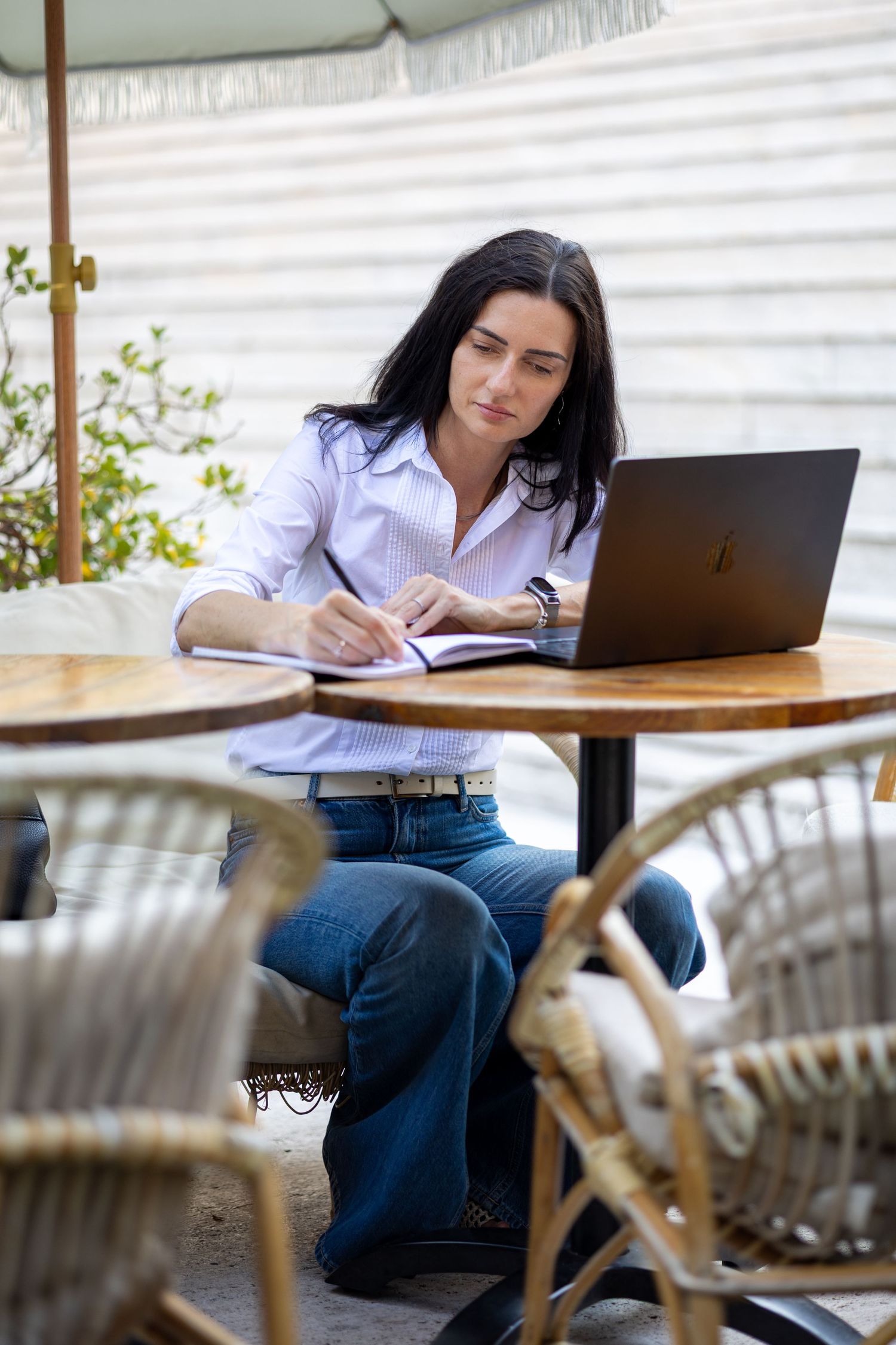 Woman in white shirt writing in notebook at outdoor cafe table with MacBook laptop.