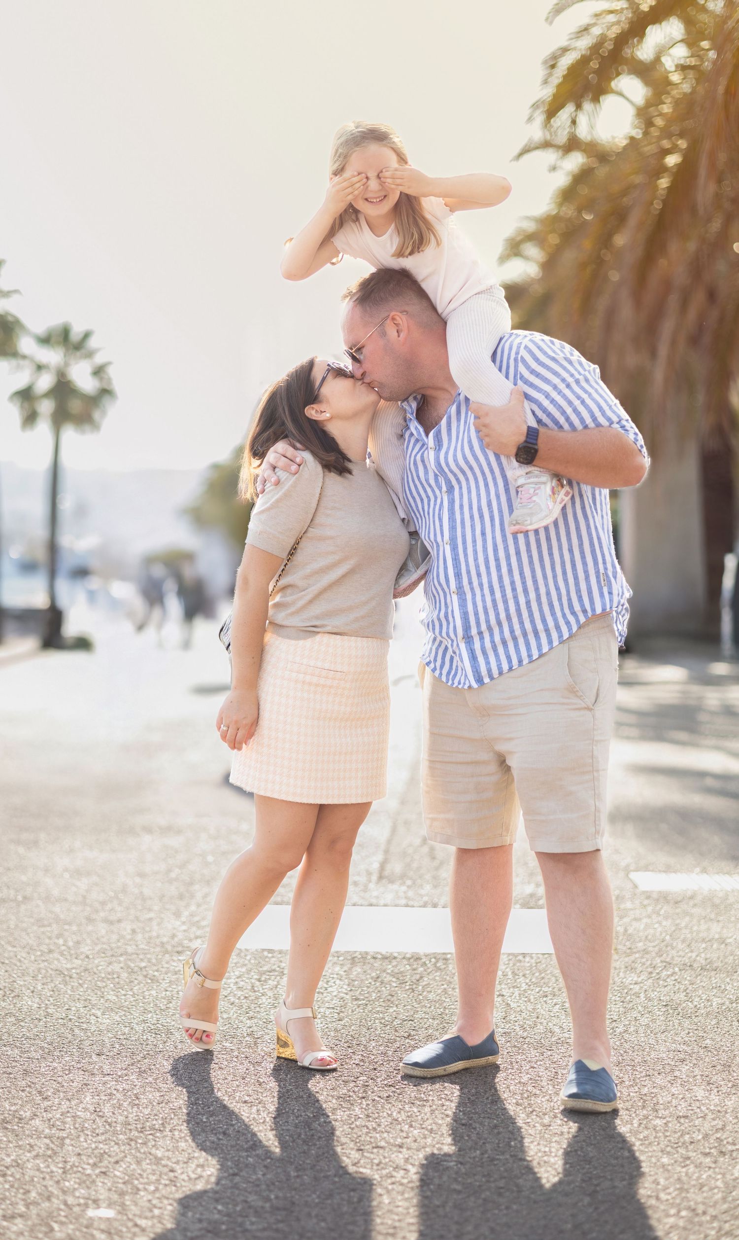 Happy family kissing outdoors on a sunny street with palm trees, child sitting on fathers shoulders.