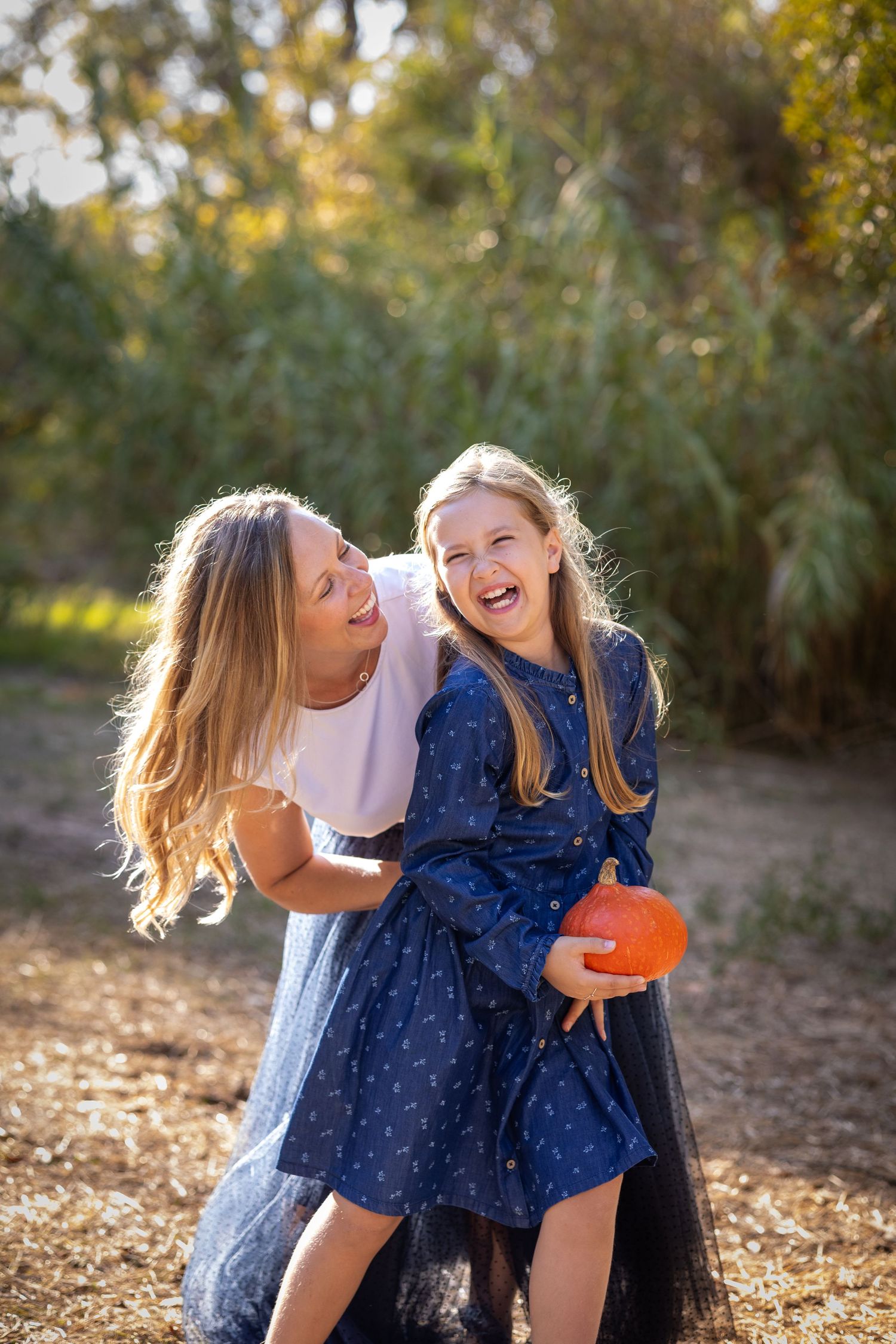 A laughing mother and daughter share a joyful moment outdoors, with the girl holding a small orange pumpkin.