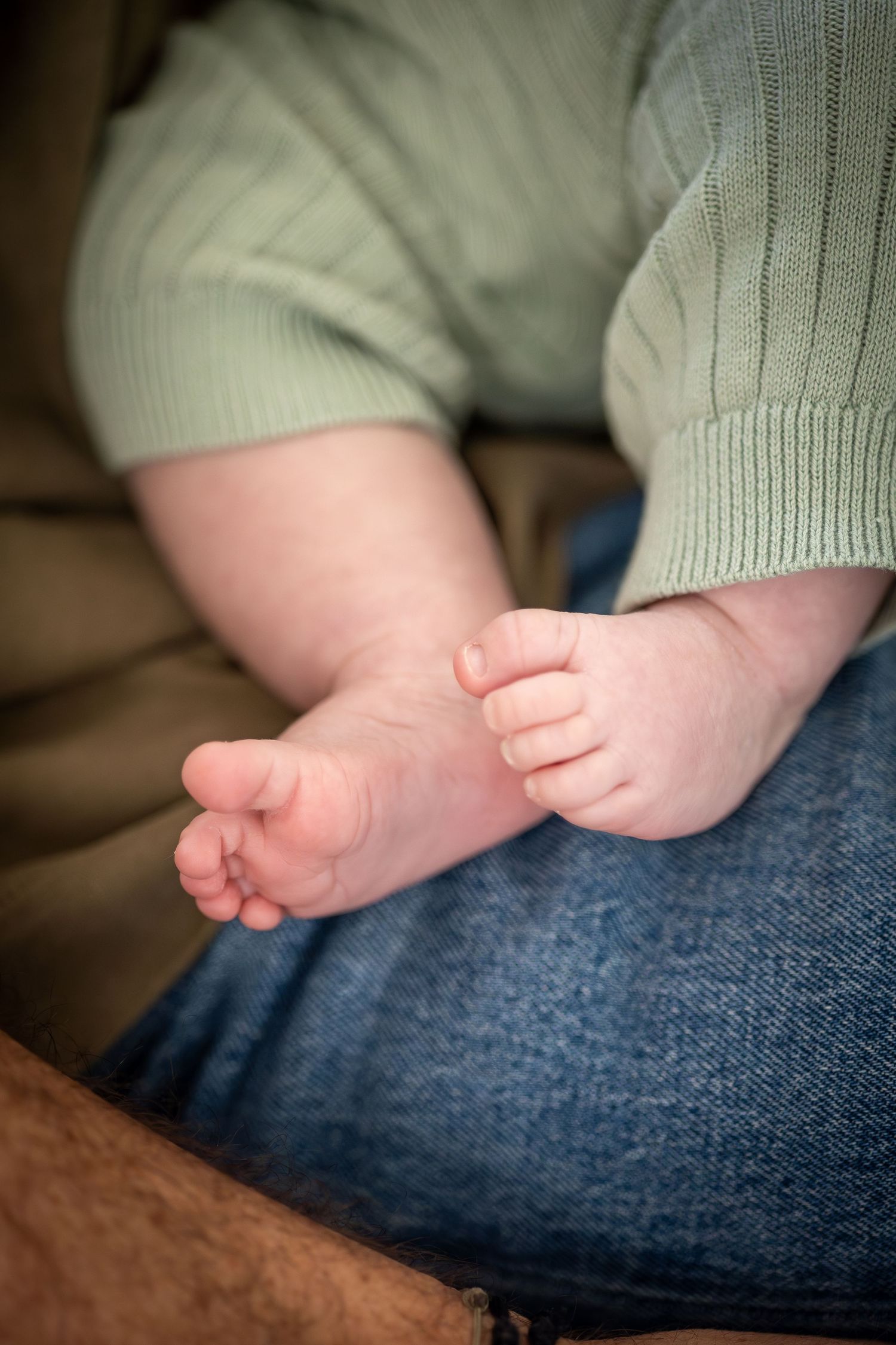 Close-up of newborn baby feet in green outfit resting on parent's denim-clad lap.
