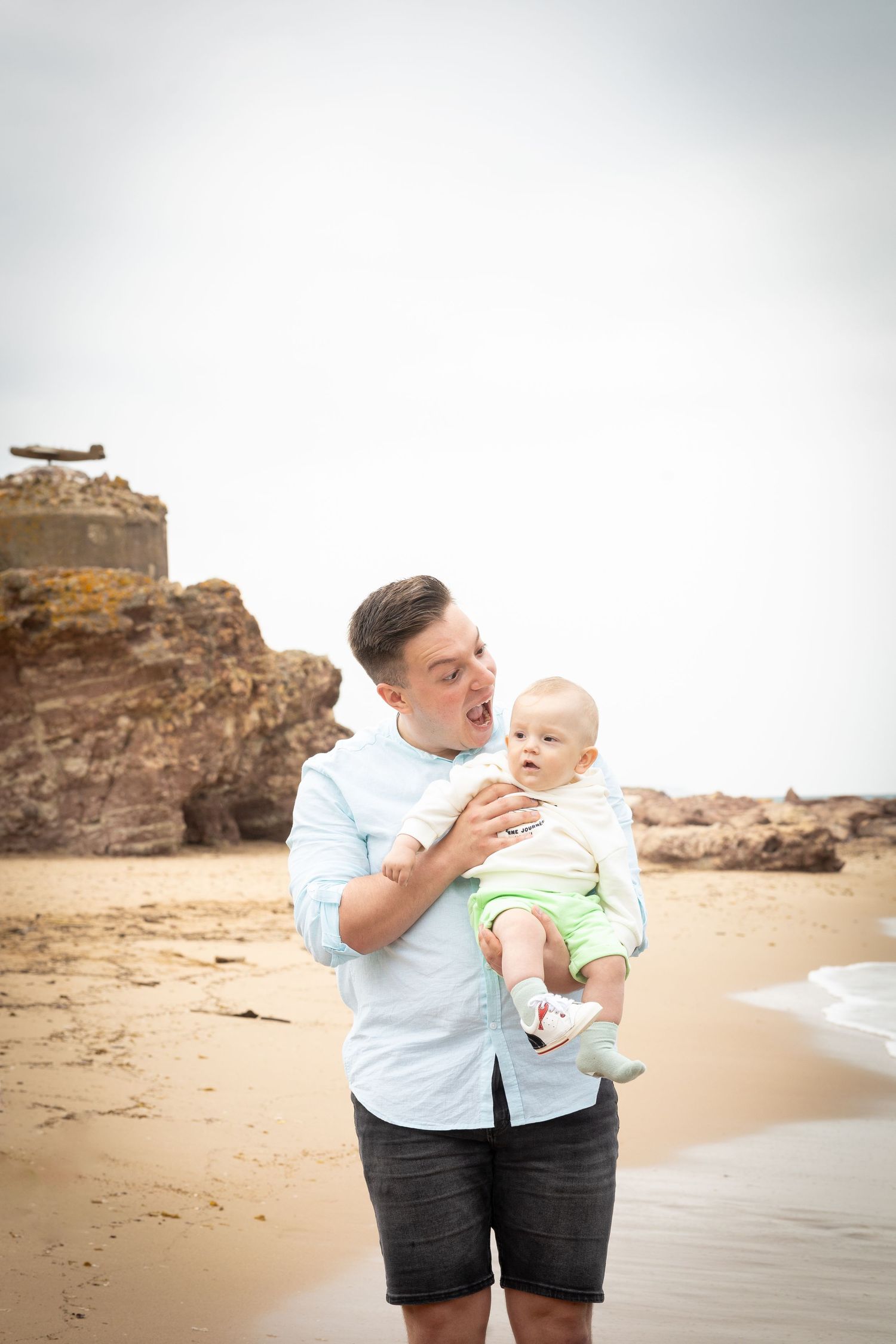 Young father making funny faces while lovingly holding his baby on a sandy beach with rocky cliffs in the background.