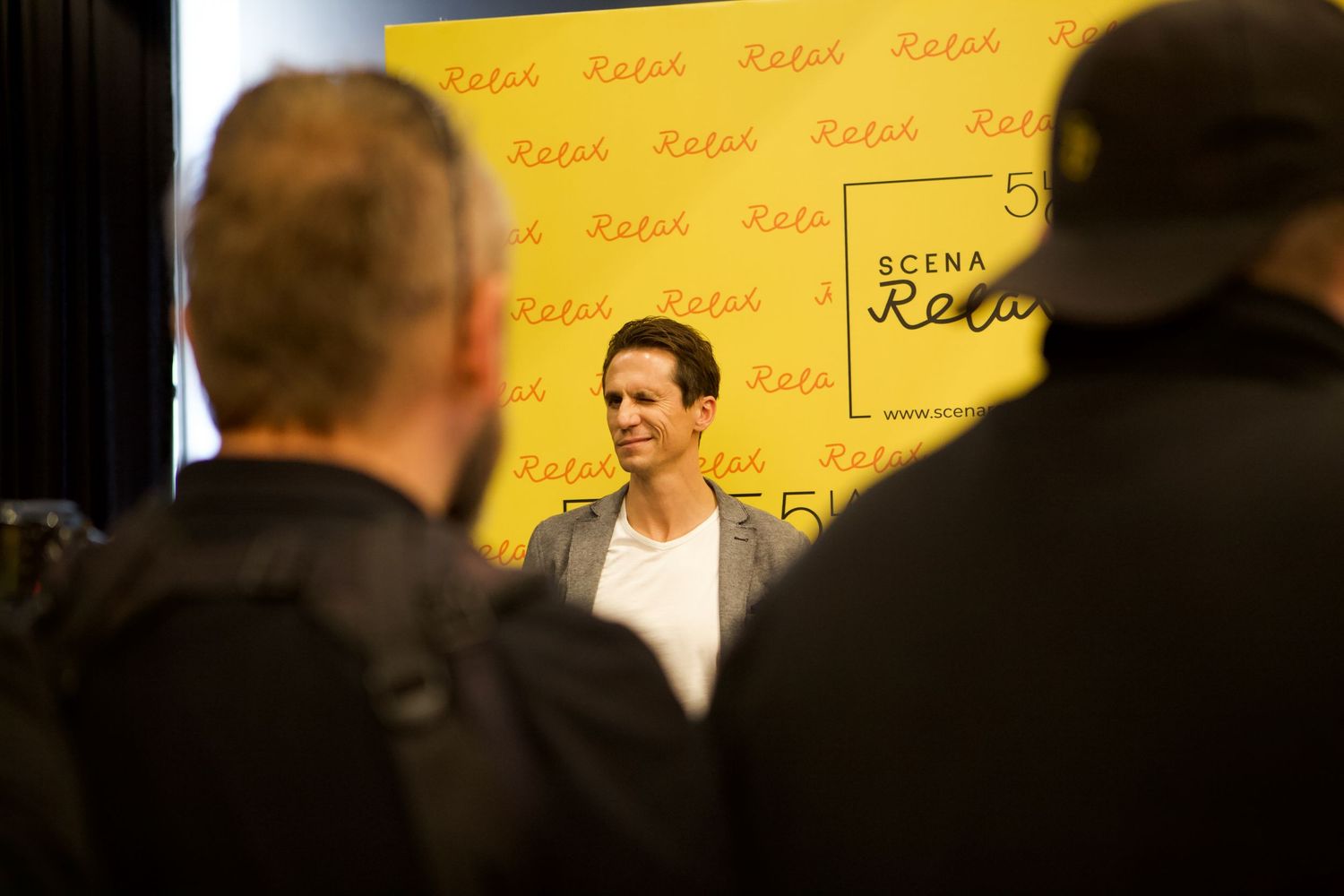 Person in a grey blazer speaking at a press event in front of a yellow branded backdrop.