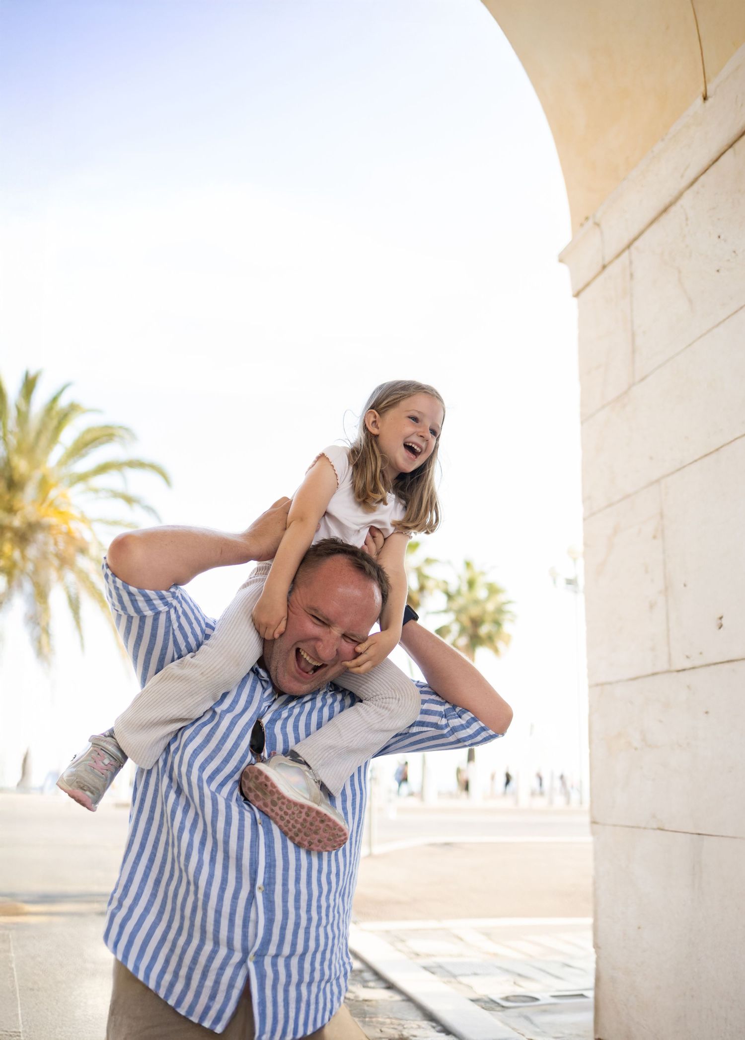 Joyful father carrying laughing daughter on his shoulders near a sunlit archway with palm trees in the background.