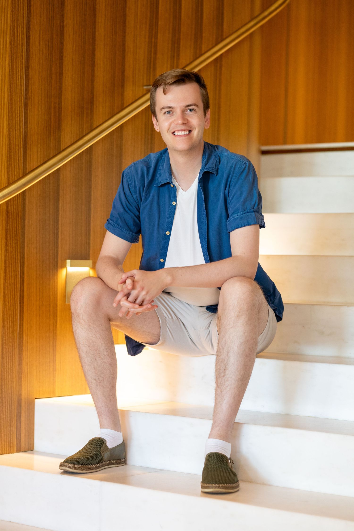Young man smiling, sitting on modern white stairs with wood paneling, wearing blue shirt and khaki shorts.