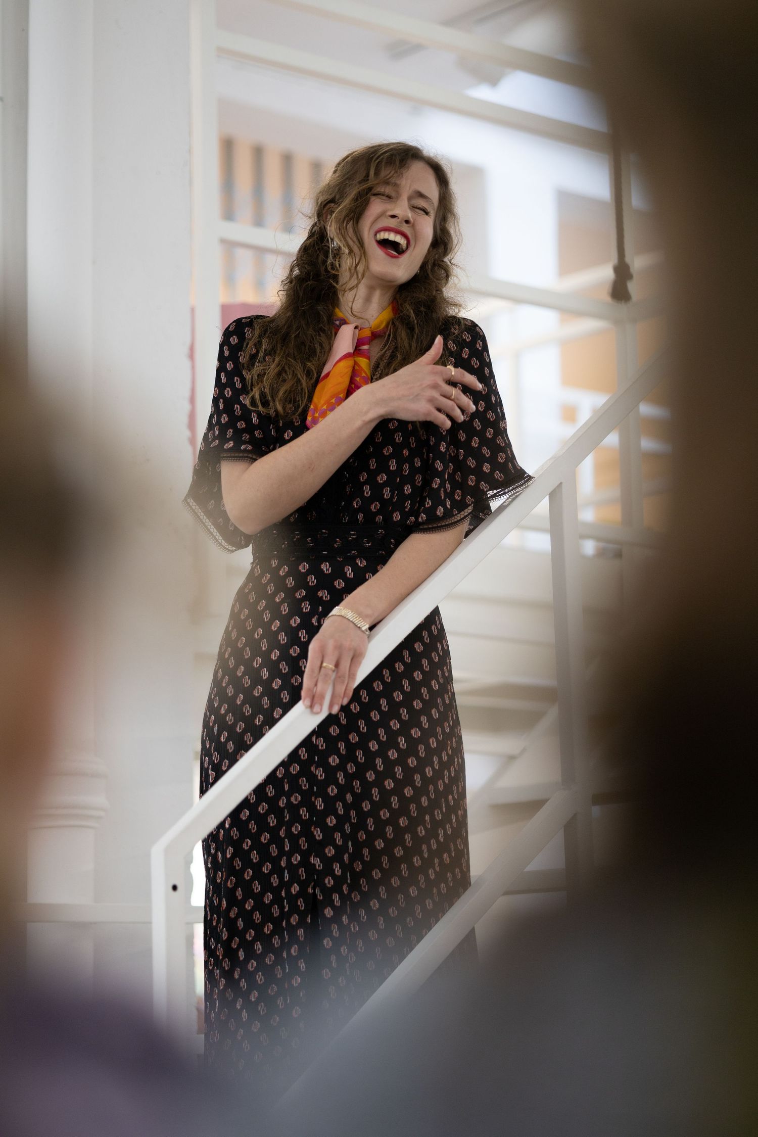 Joyful woman in black patterned dress laughing on white staircase at art gallery.