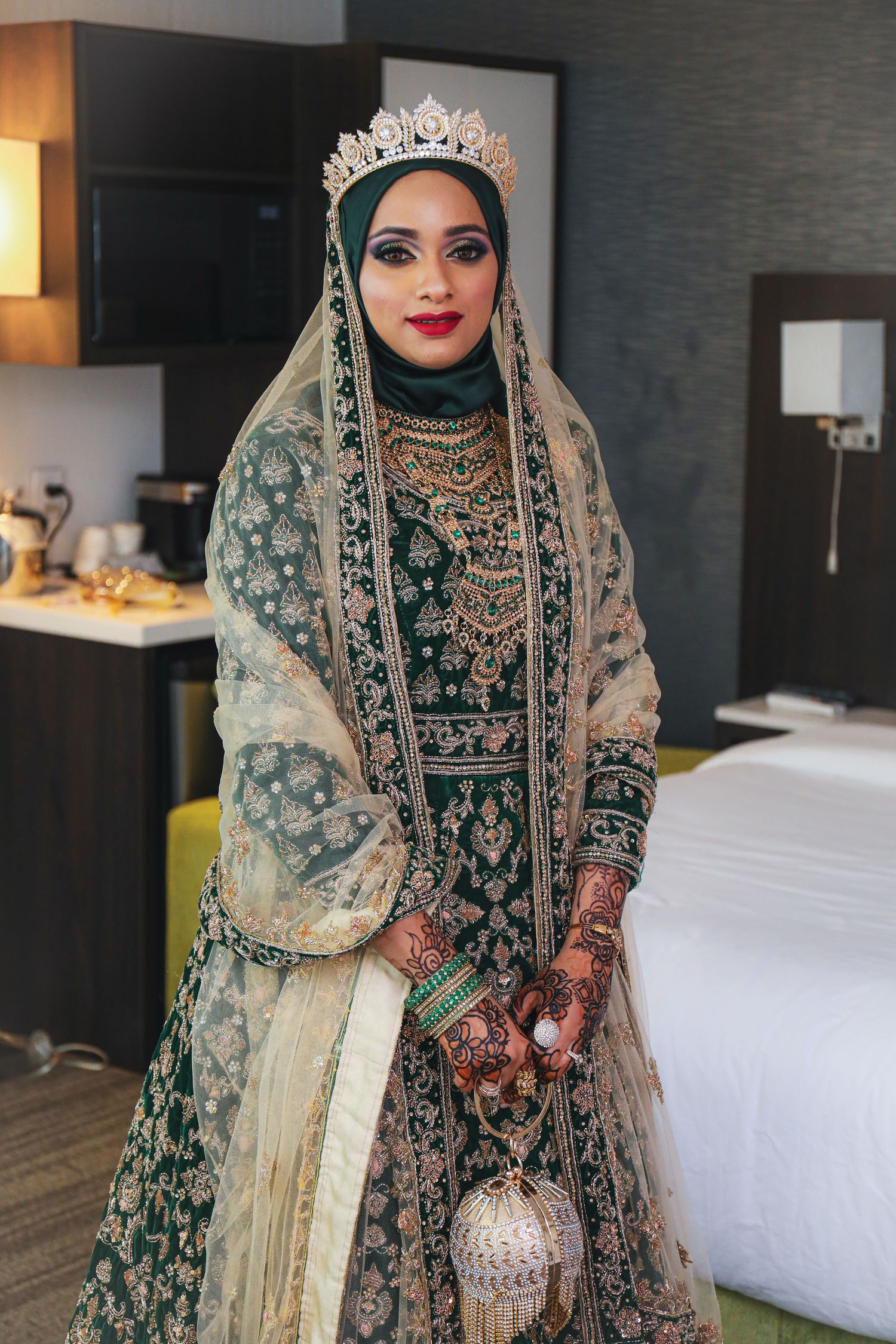 Bride in emerald and gold traditional dress with crown and metallic clutch standing in hotel room.