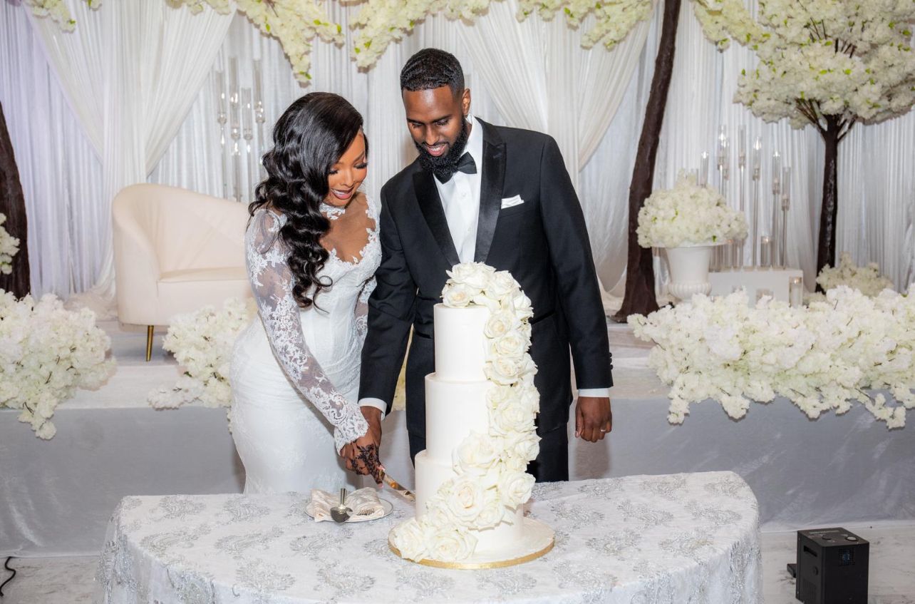 Elegant wedding cake cutting scene with an ornate white tiered cake against a floral backdrop.