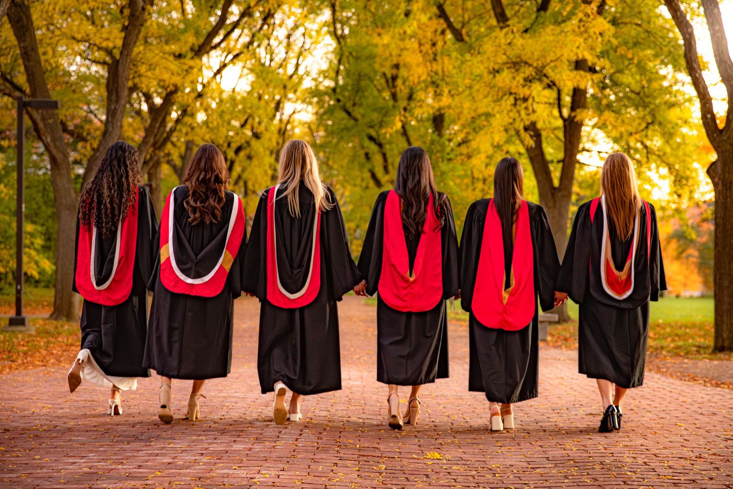 Group wearing black dresses and red shoes walks along a tree-lined path during golden autumn sunset.