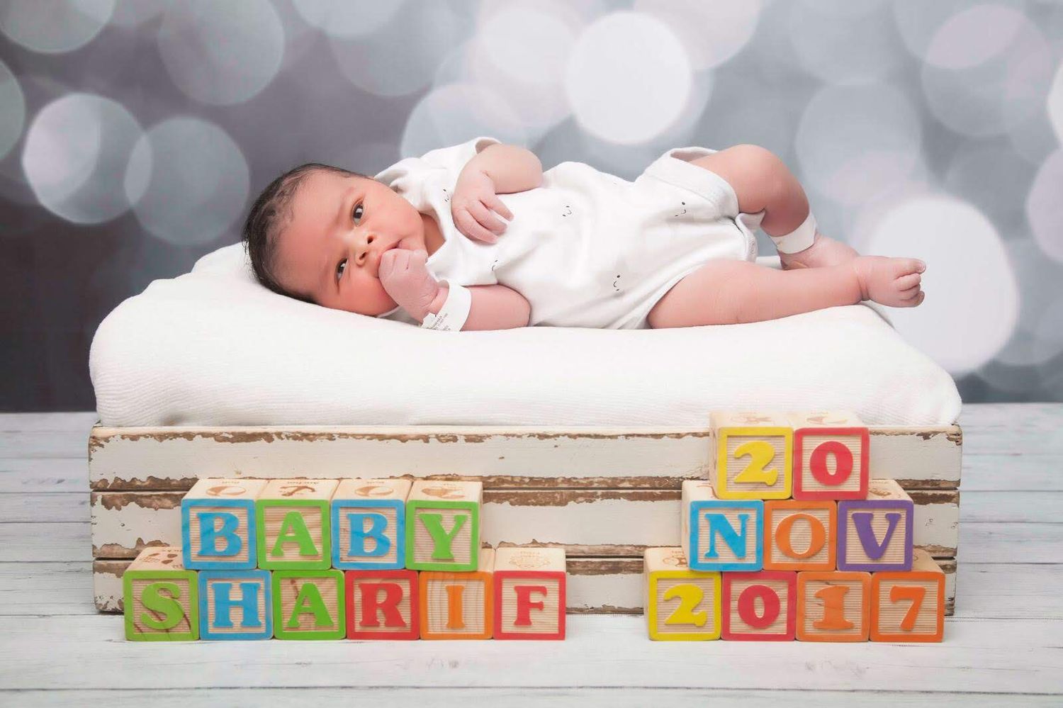 Baby lying on white surface with wooden blocks displaying date and name 'BABY SHARIF 20 NOV 2017'.