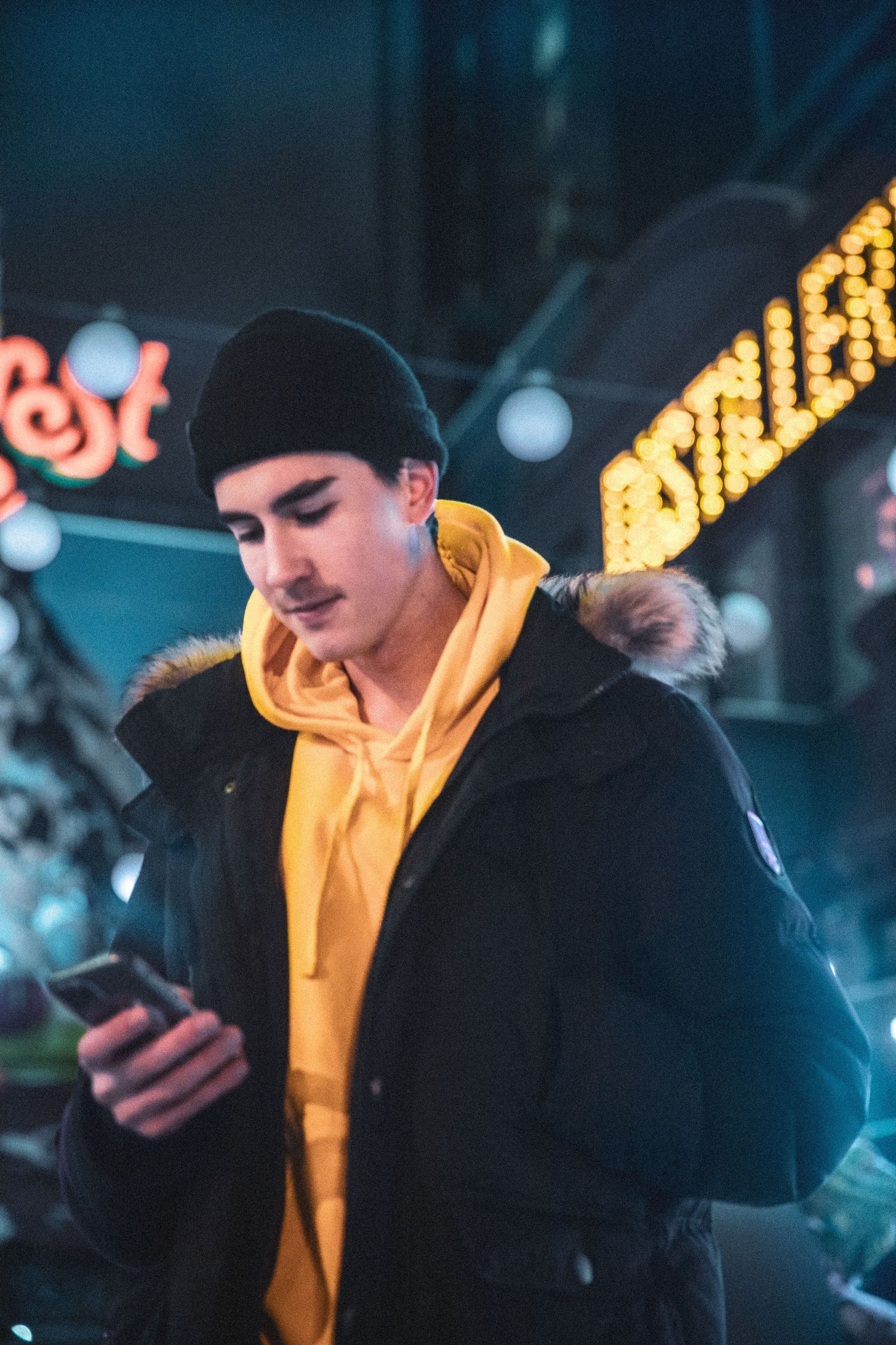 Young man in yellow hoodie and winter jacket looking at phone with holiday lights in background.