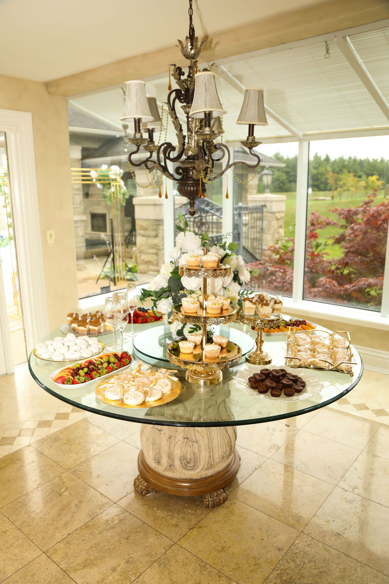 Elegant dessert display on glass table with ornate chandelier and garden view.