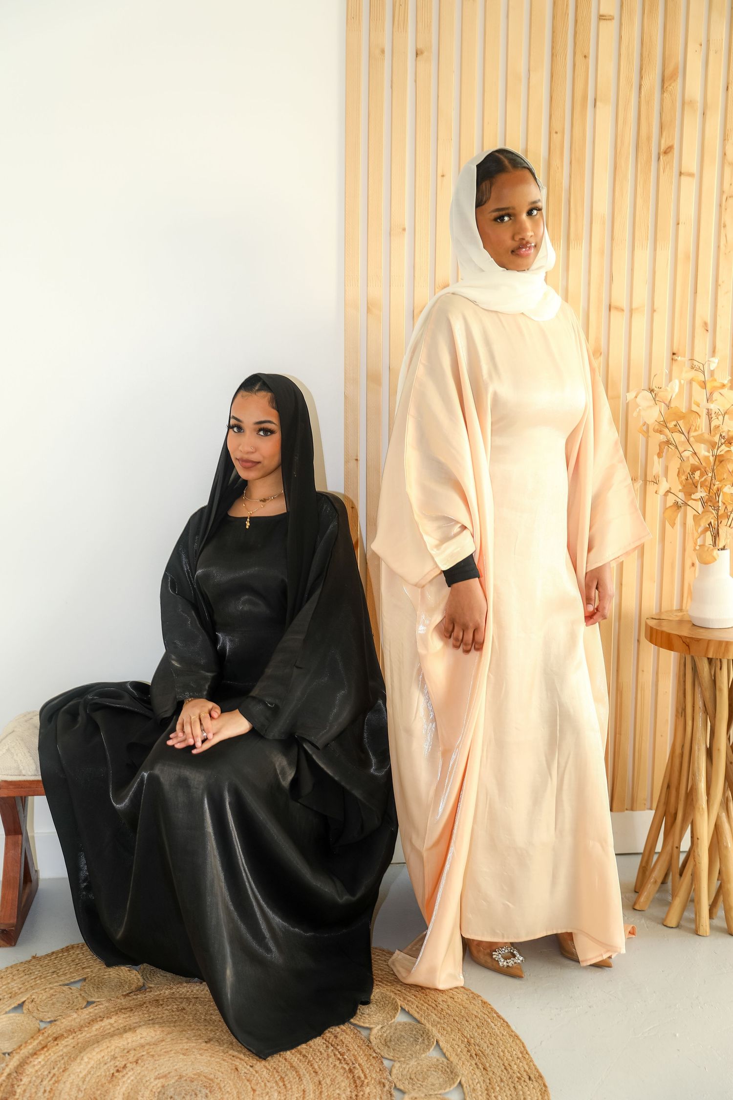 Two women in black and white abayas posing against wooden panel wall.