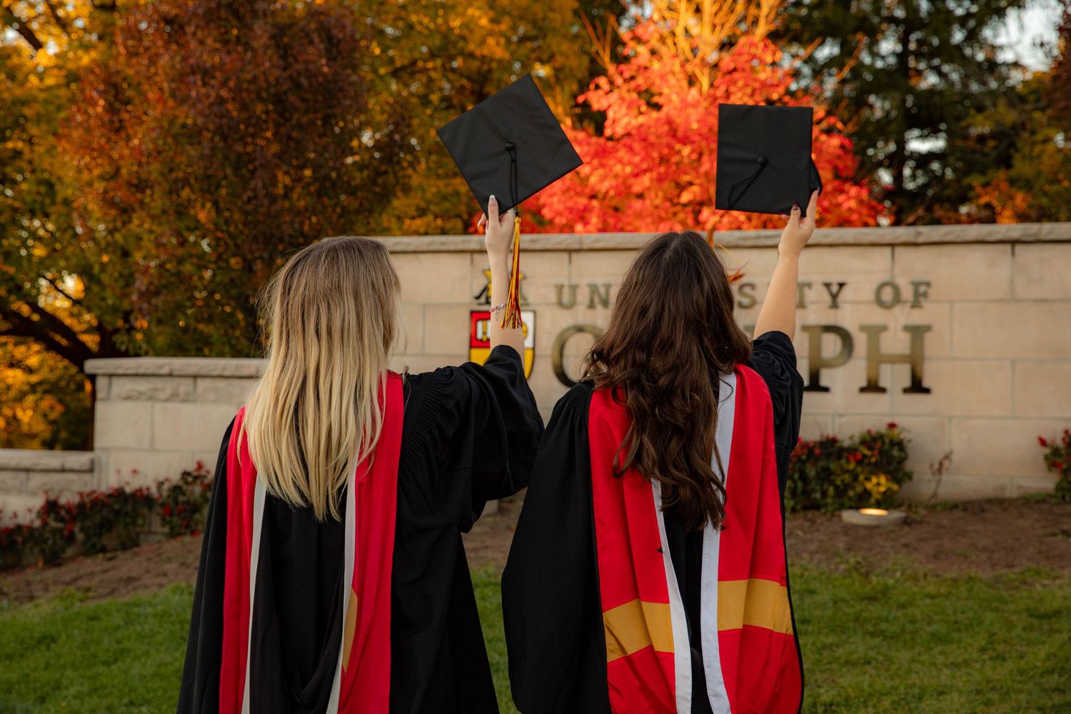 A group in red and black graduation robes stands together outdoors during autumn with colorful fall foliage in the background.