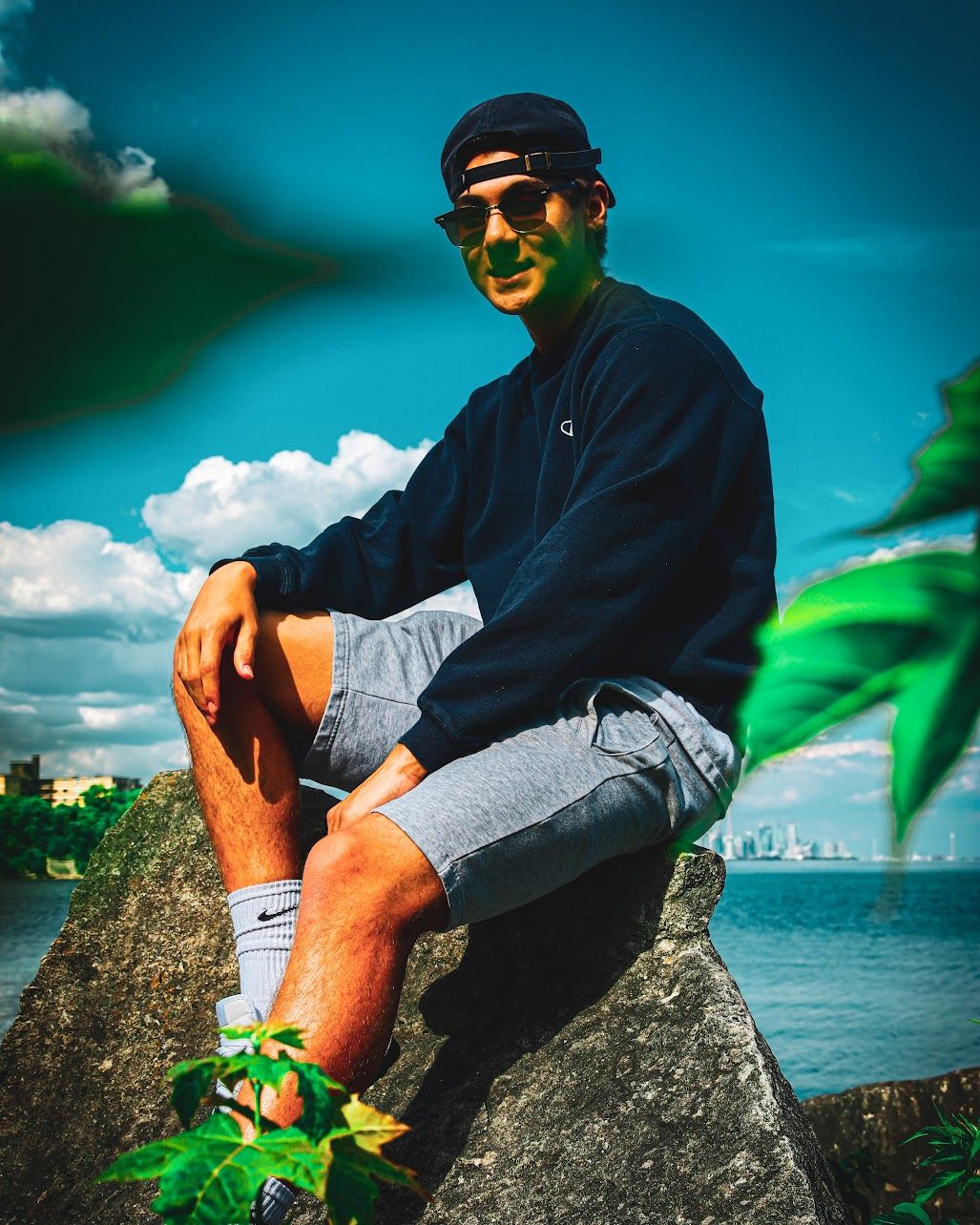 Person sitting on rocky outcrop overlooking water wearing sunglasses and casual attire.