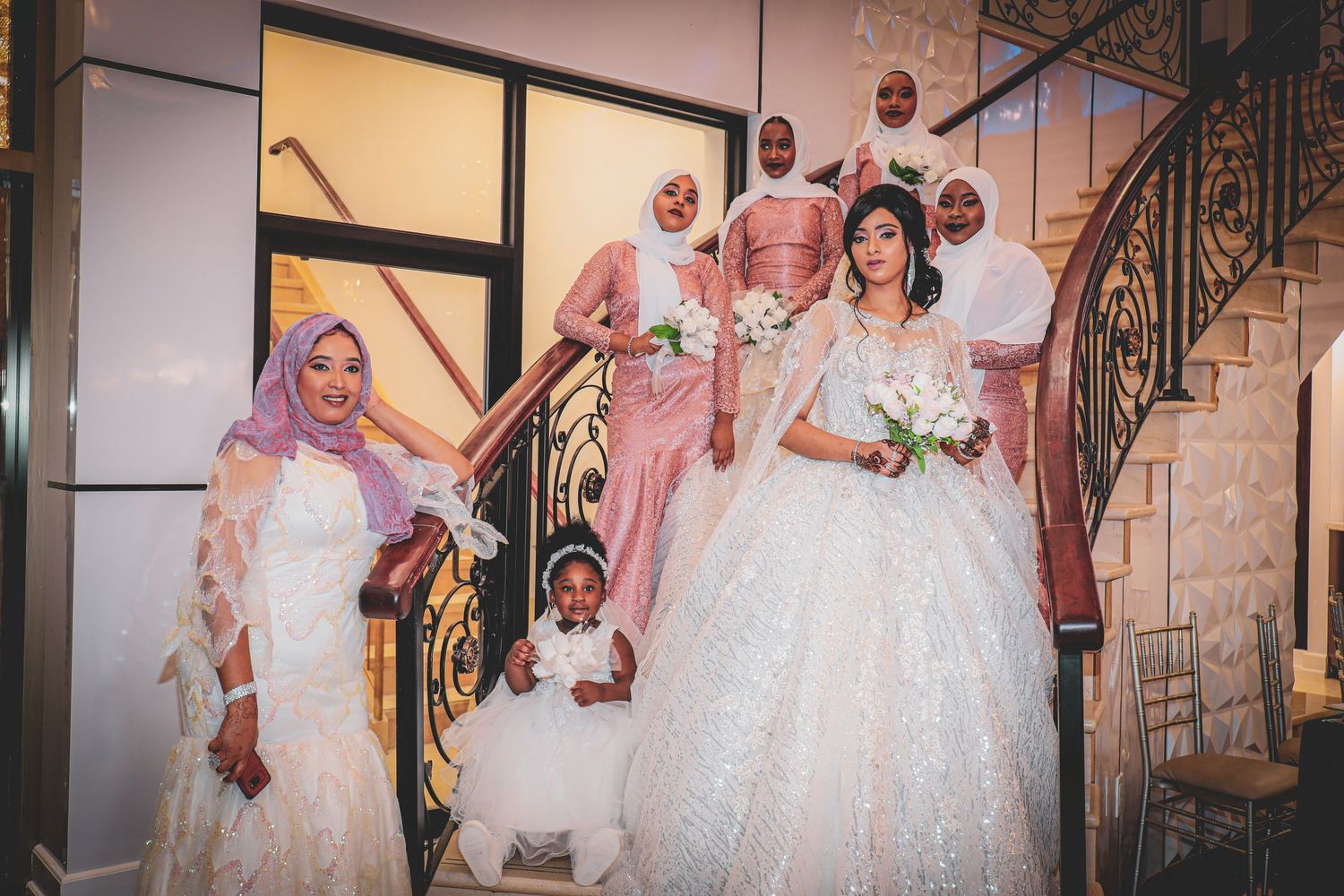 Wedding party posing on elegant staircase with bride in white dress and family members.