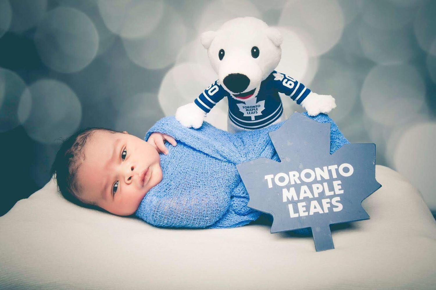 Newborn baby wearing Toronto Maple Leafs outfit with team mascot plush toy on sparkly background.