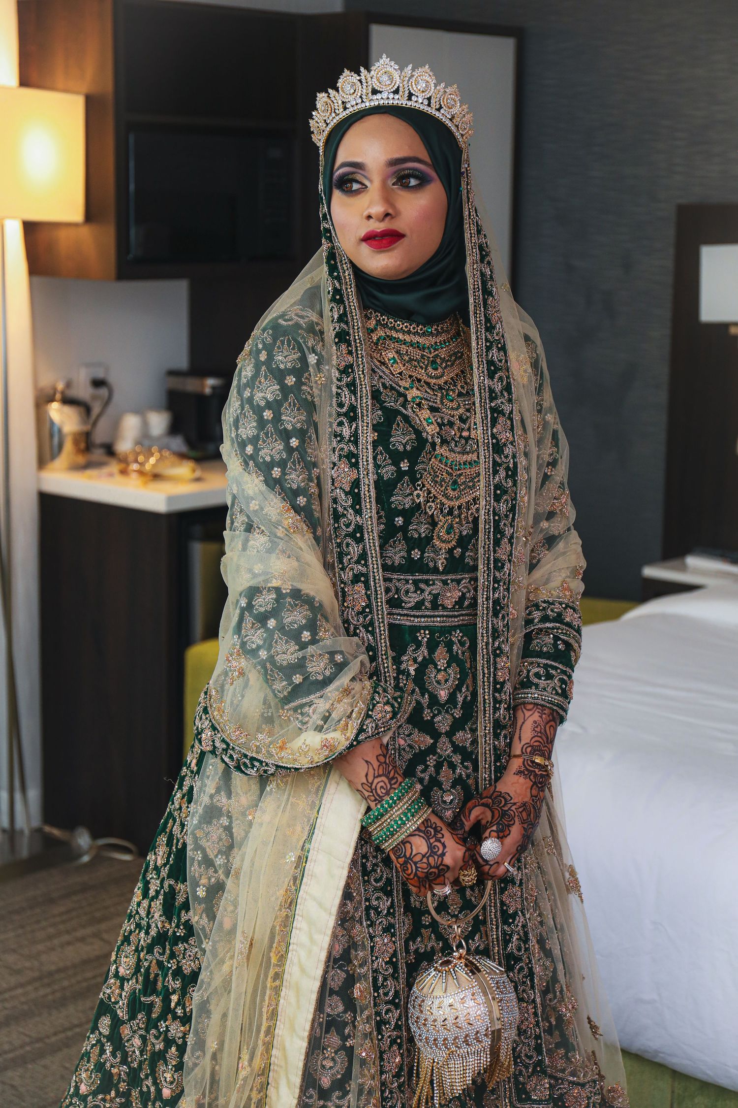 Muslim bride wearing an ornate emerald green and gold lehenga with crown and intricate henna designs.