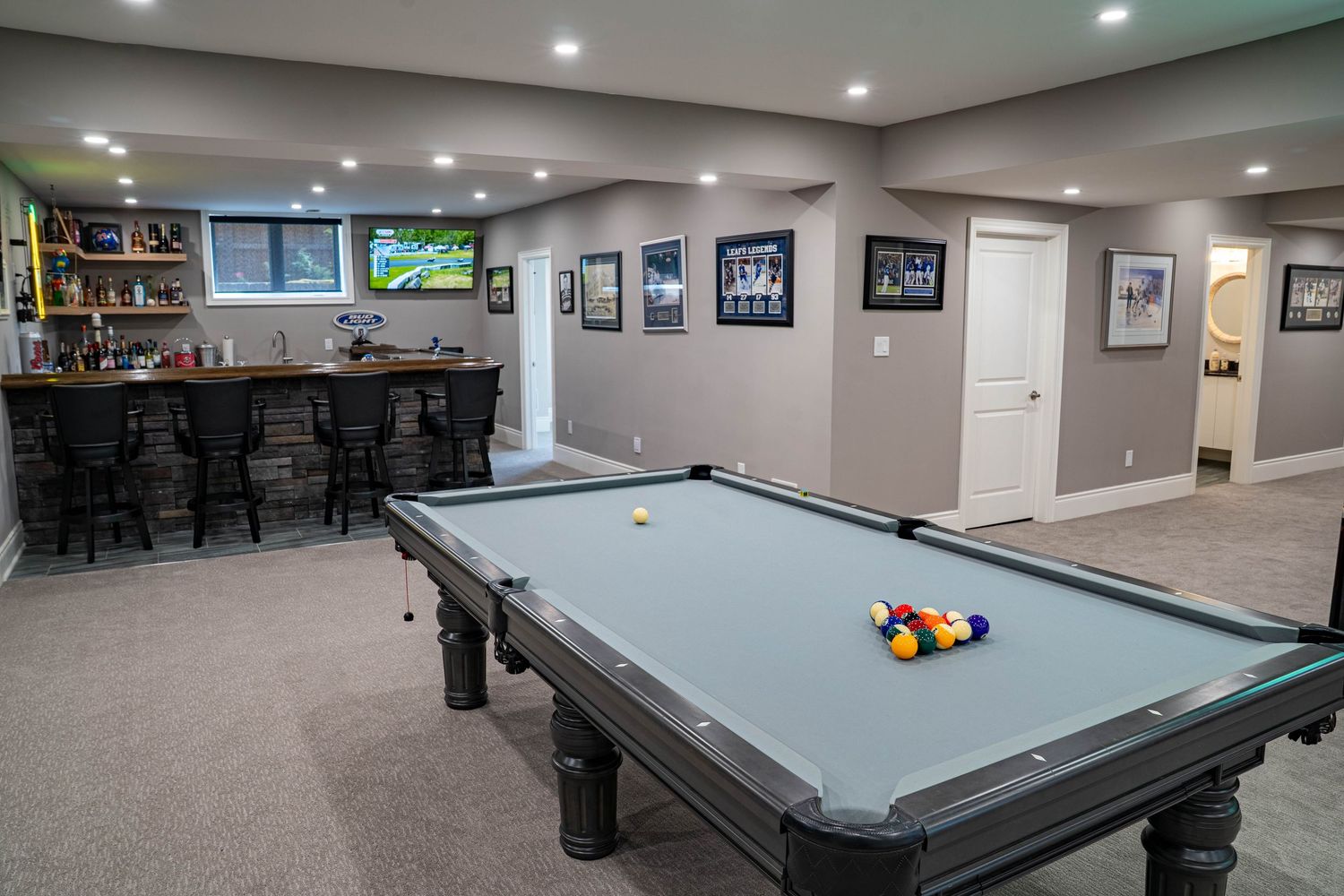 A basement game room features multiple pool tables with blue felt under recessed lighting and framed photos on the walls.