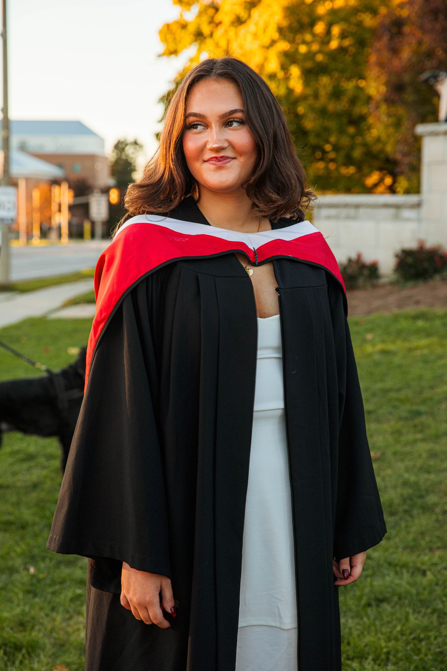 Graduate in black academic regalia and white dress stands outdoors during golden hour sunset.