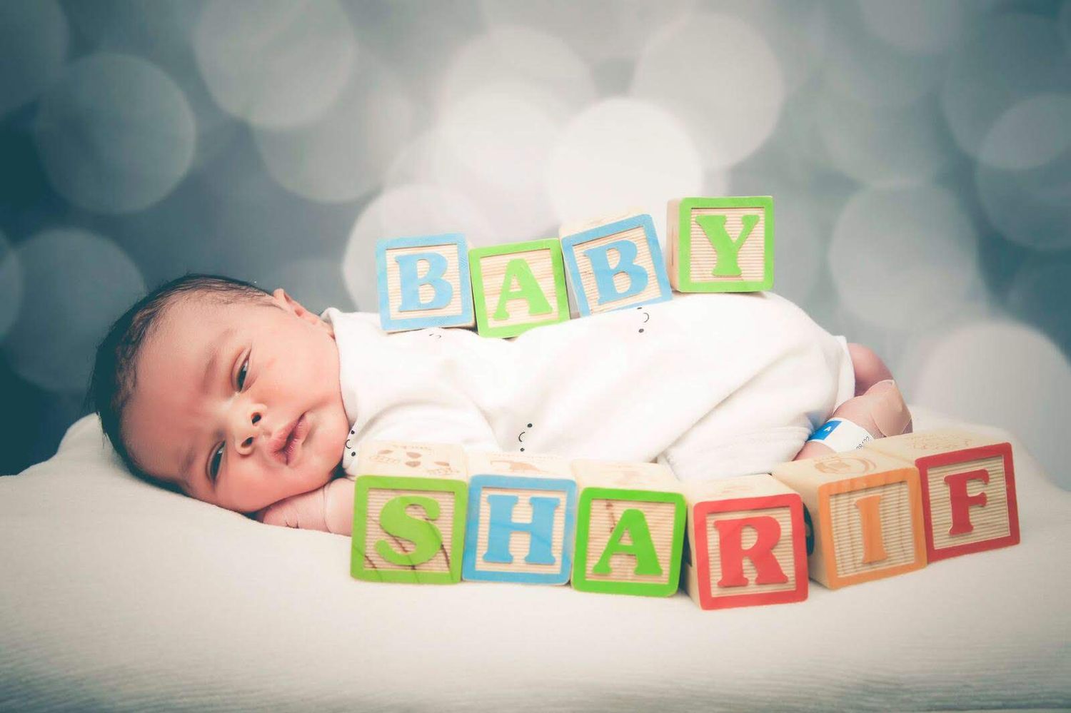 Newborn baby lying on soft white fabric with colorful alphabet blocks spelling 'BABY SHARIF' against bokeh background.