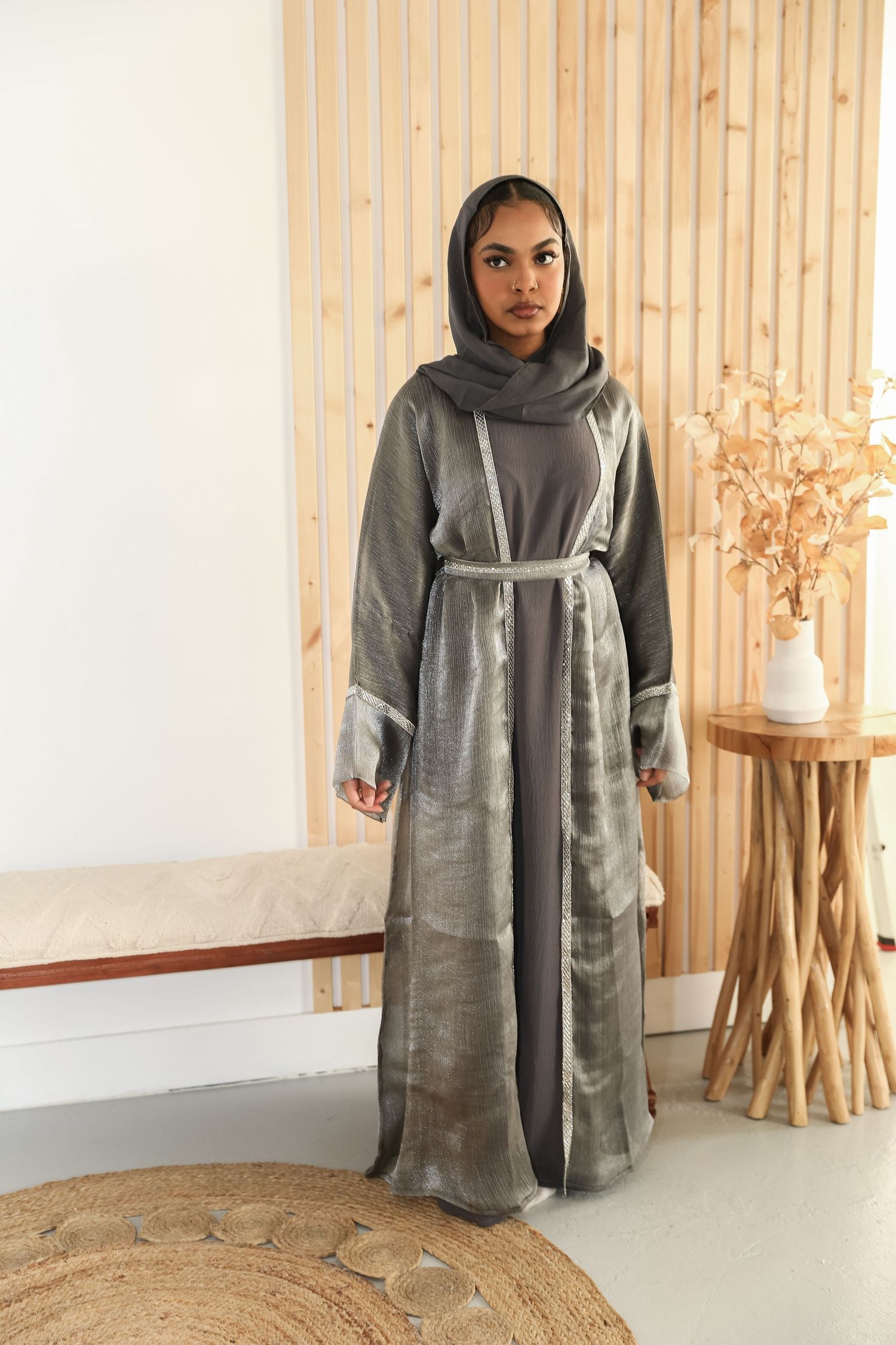 Woman wearing grey belted abaya and hijab standing against wooden wall.