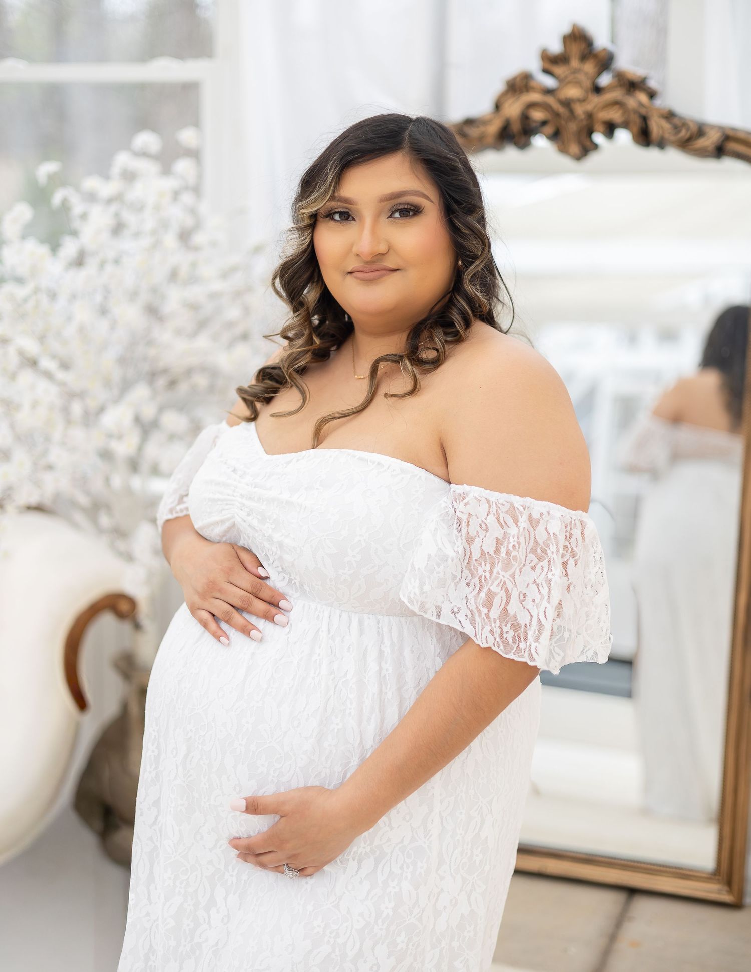 Maternity photoshoot featuring an elegant white lace off-shoulder gown in a bright indoor setting.