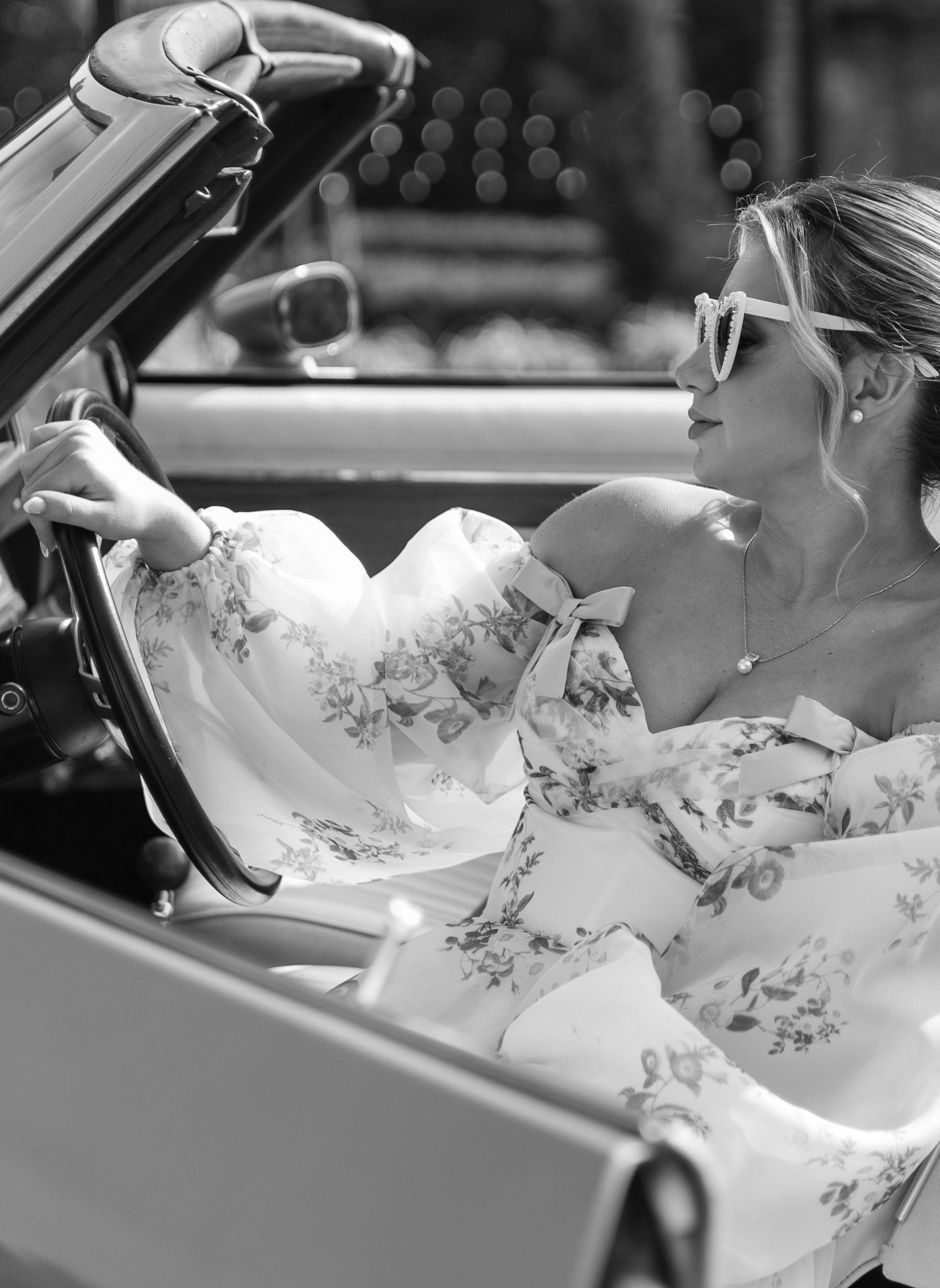 Black and white photo of a bride's dress flowing out of a classic car in an elegant wedding moment.