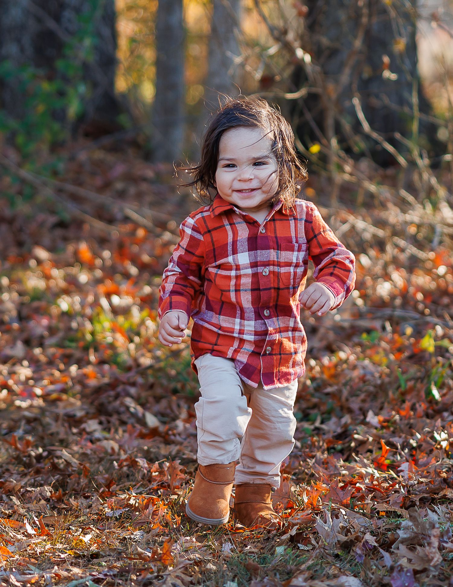 Small child wearing red plaid shirt and khaki pants plays in autumn leaves on forest ground during golden hour.
