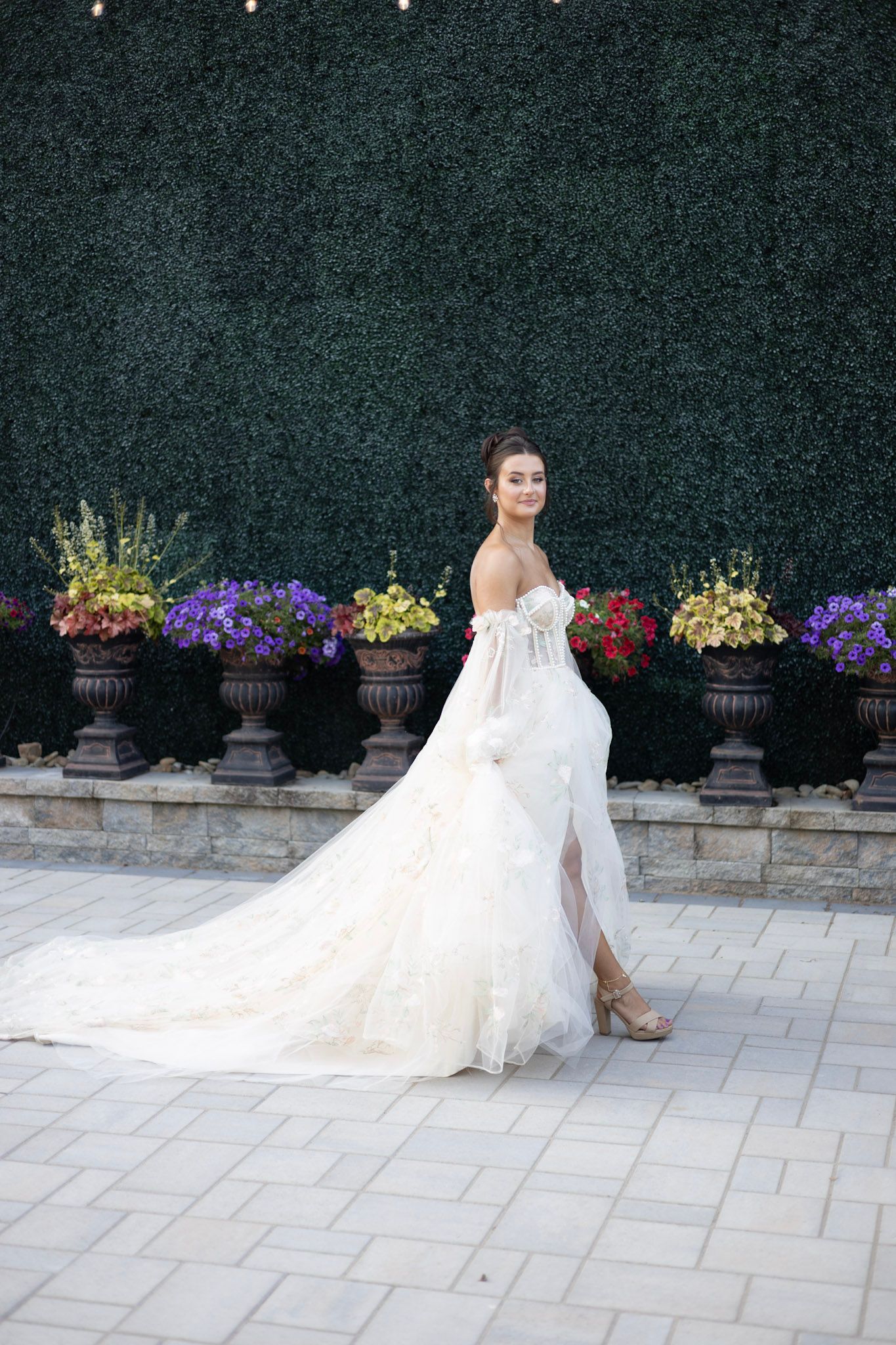 Elegant wedding dress with flowing train photographed against stone patio and colorful garden flowers.