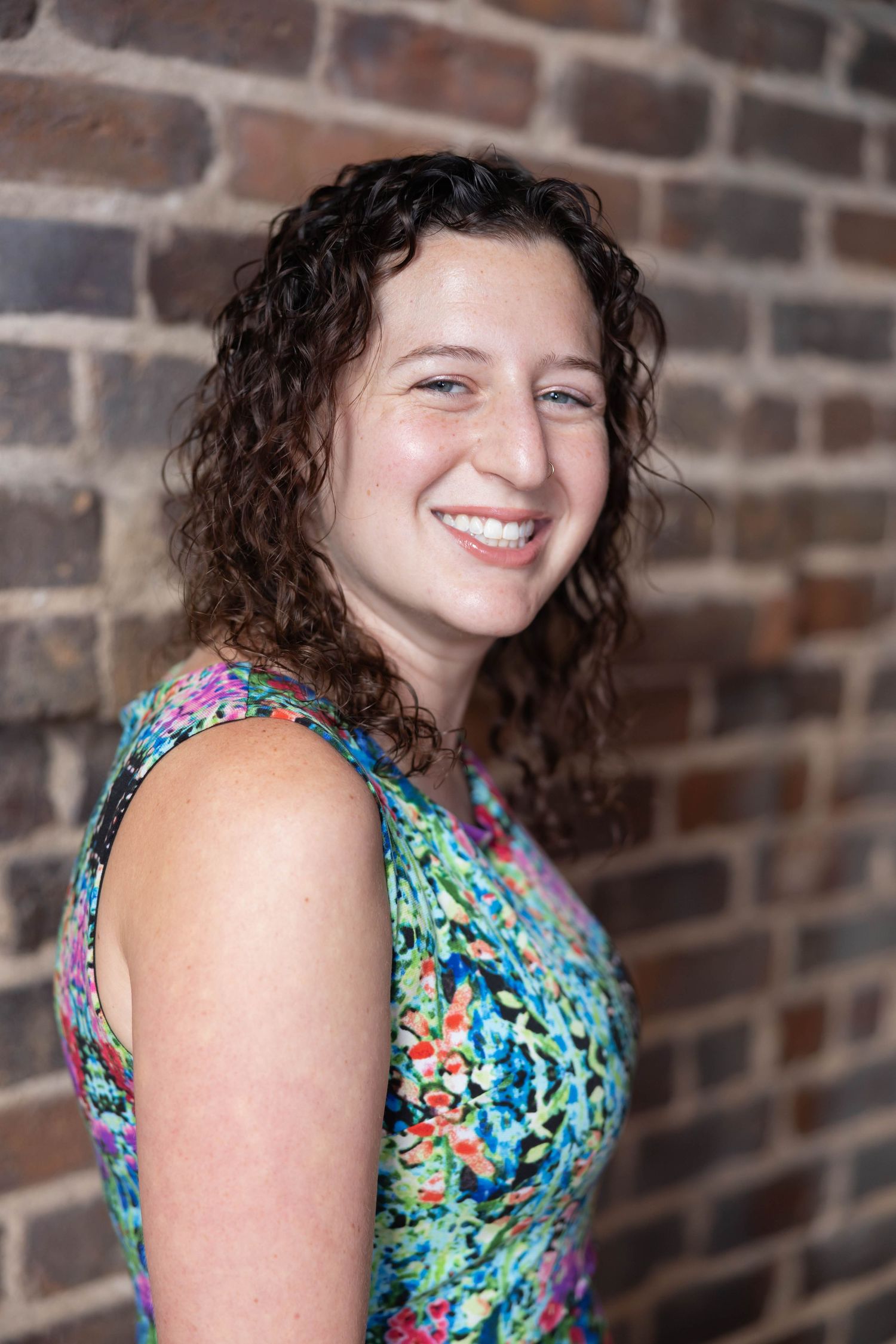 A person in a colorful floral dress poses against a brick wall with a warm smile and curly hair.