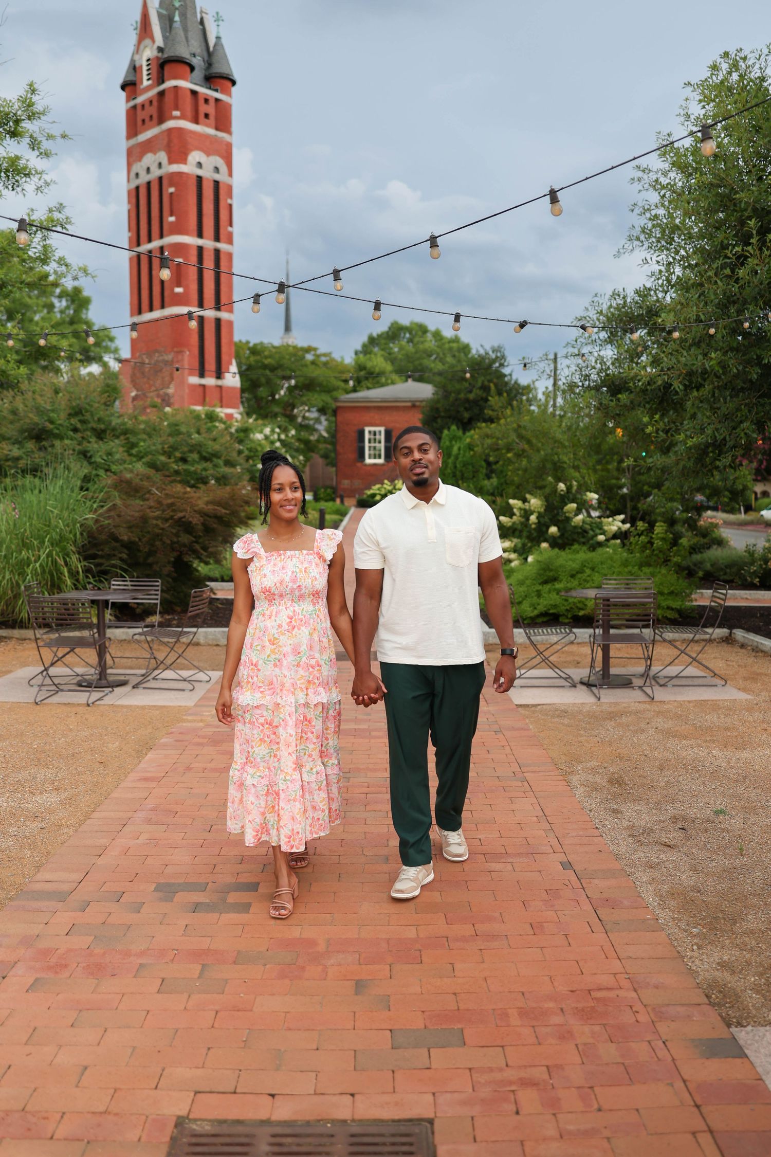 A couple strolls on a brick walkway with a tall red brick clock tower and string lights visible in the background.