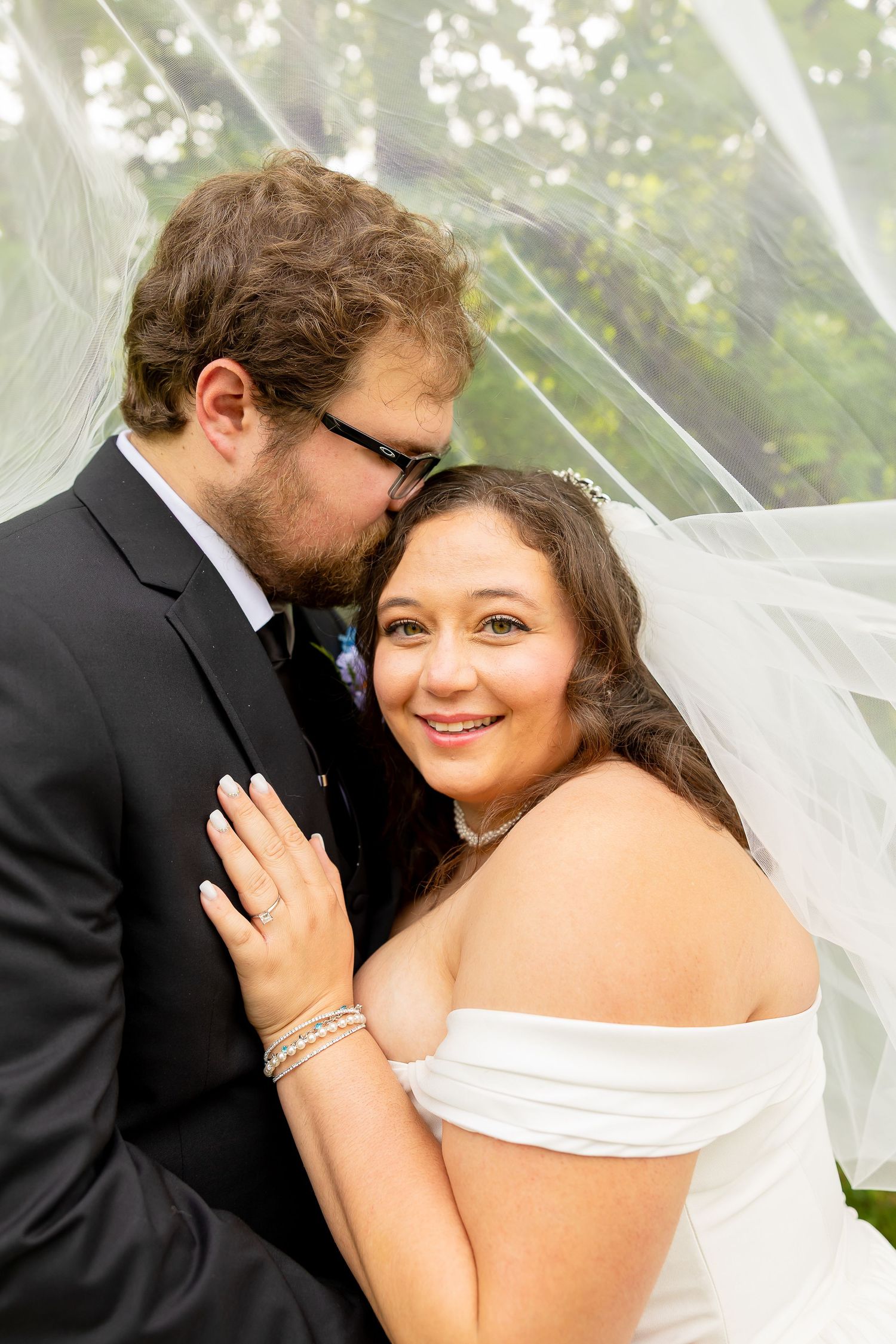 Bride in white off-shoulder gown smiles under veil as groom in black suit kisses her cheek outdoors.
