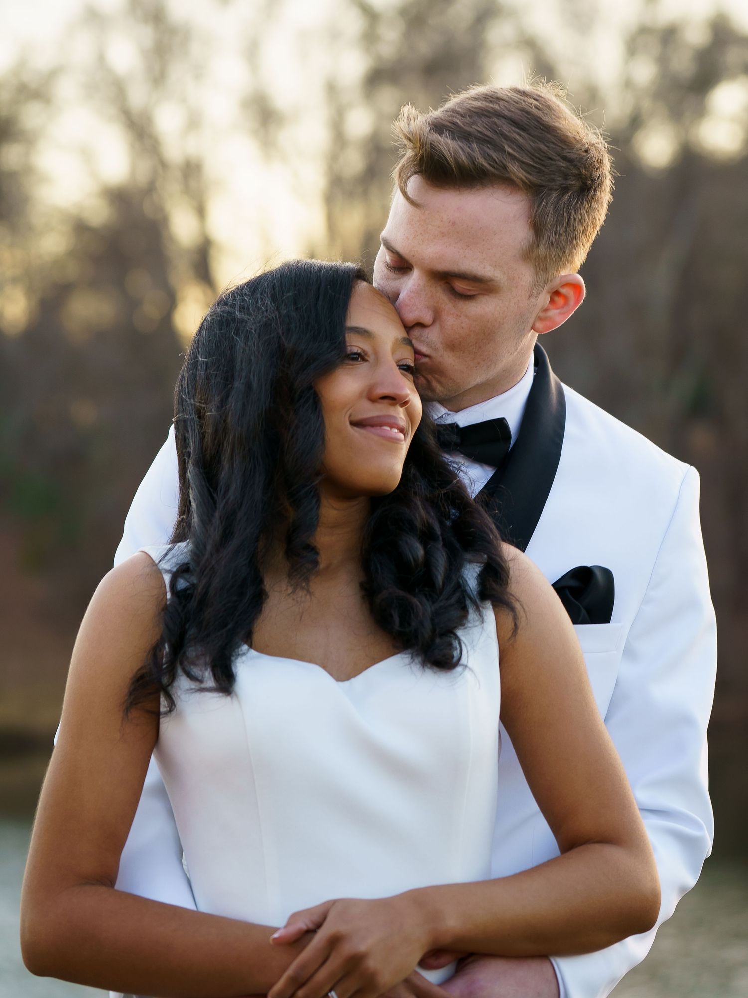 Groom in white tuxedo kissing bride on the cheek during outdoor wedding portrait.