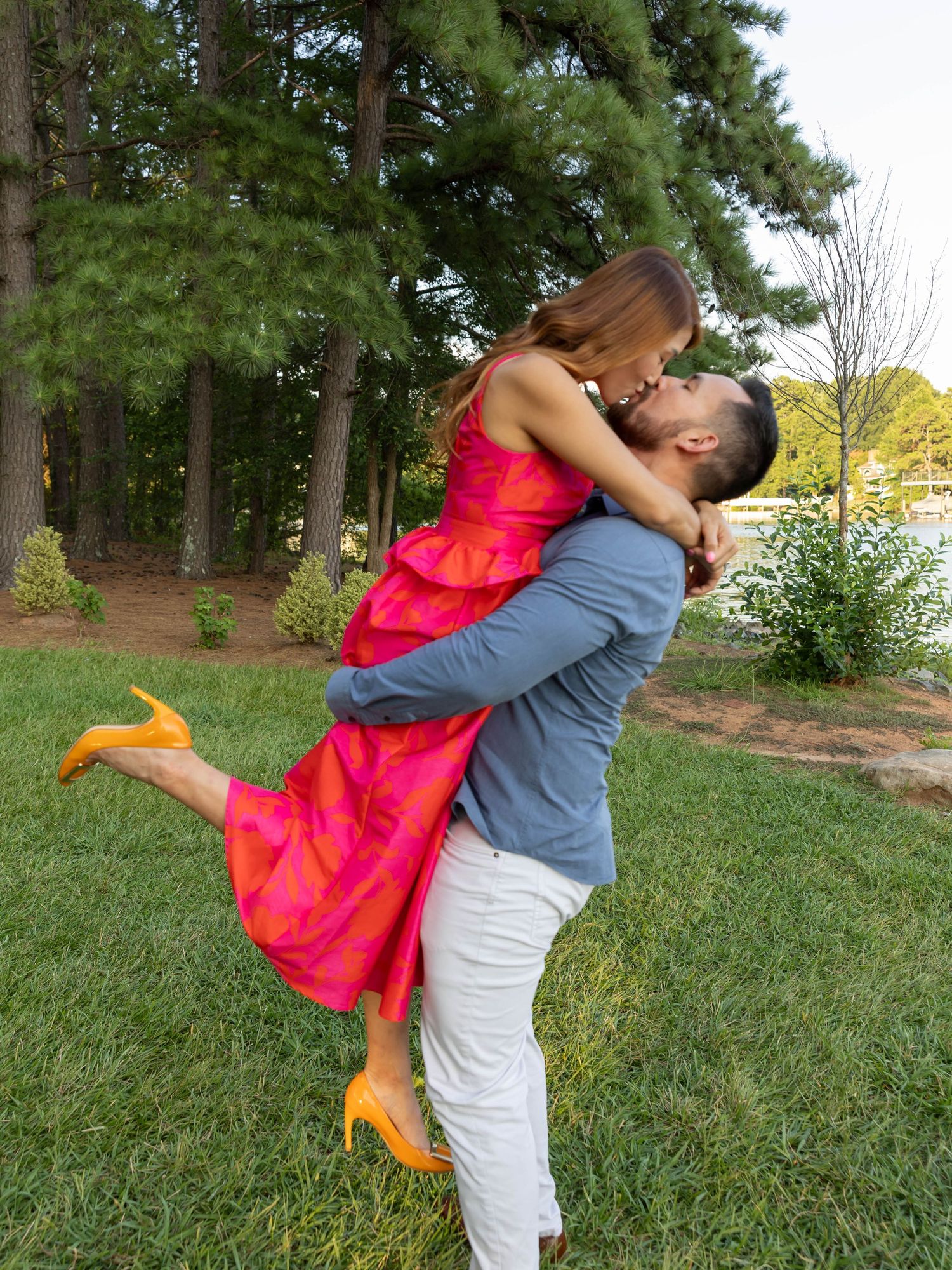 A couple shares a romantic embrace in a lush outdoor park setting, with one partner wearing a flowing bright coral dress.