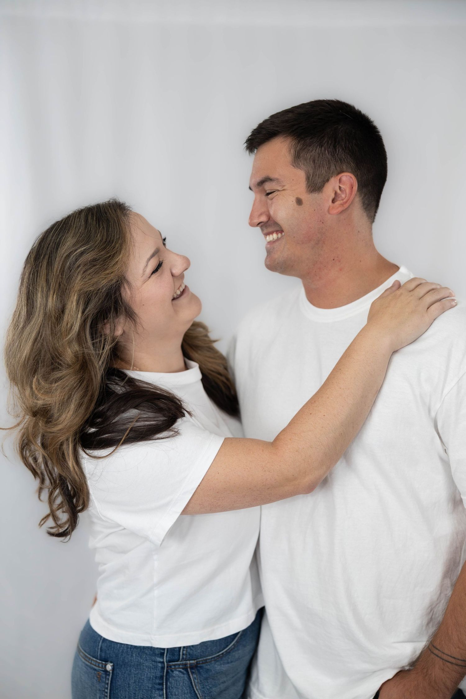 A couple wearing white t-shirts laughs together against a light background in an intimate portrait.