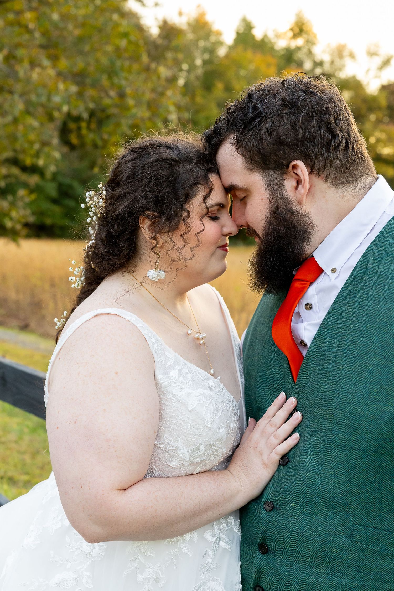 Bride and groom touching foreheads tenderly outdoors at golden hour, she in white lace gown, he in green vest with red tie.
