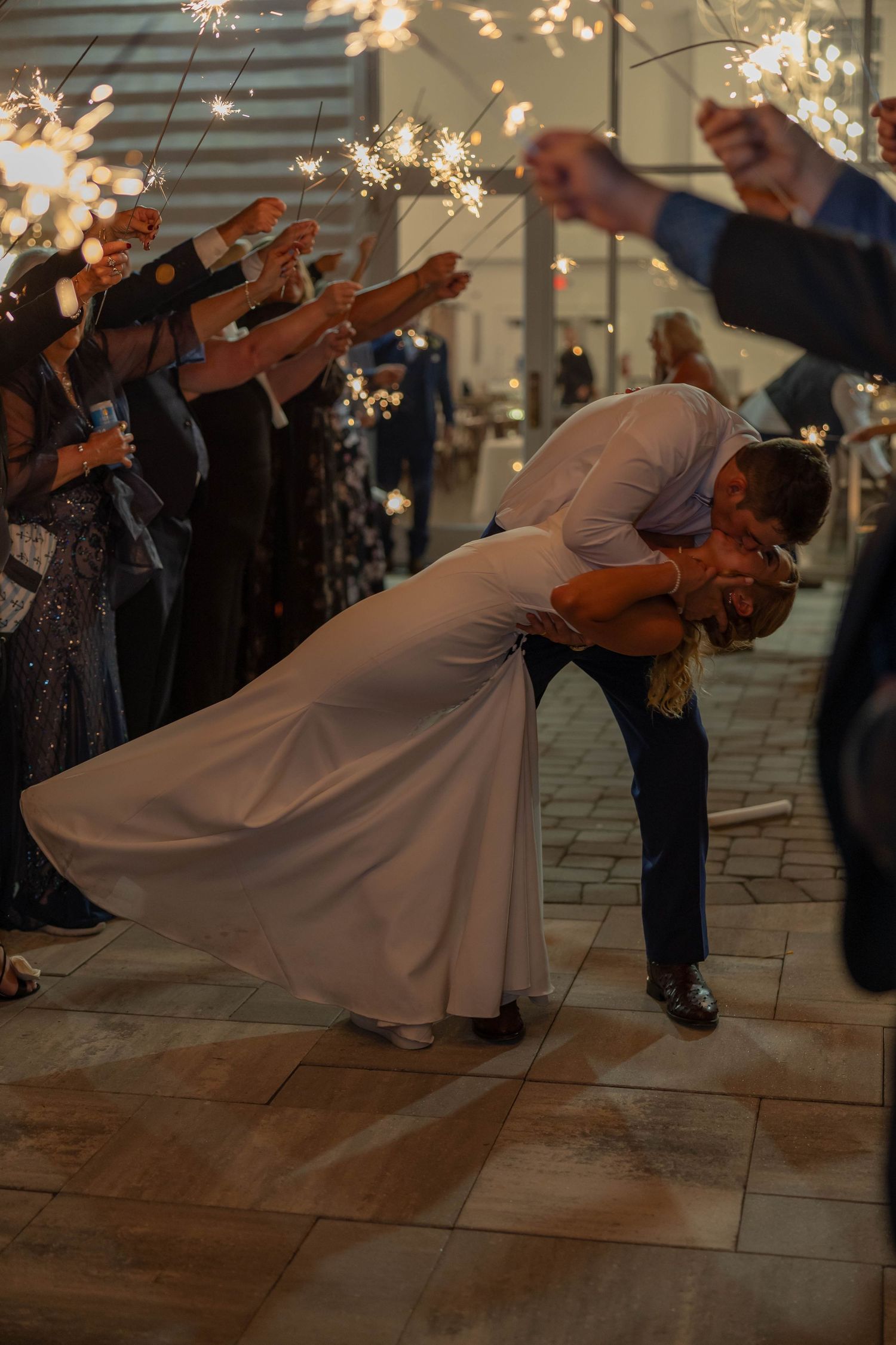 Bride and groom share a dip kiss during sparkler send-off exit at evening wedding reception celebration.
